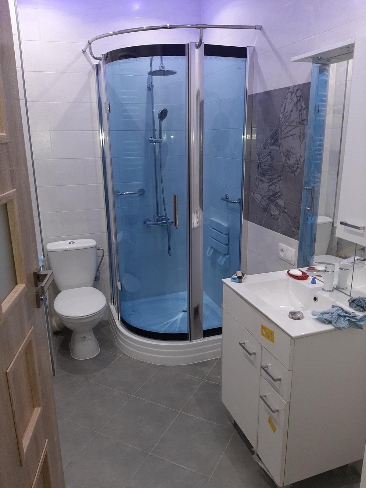 Nowoczesna łazienka po remoncie: kabina prysznicowa z hydromasażem, umywalka z szafką, toaleta. Szare płytki podłogowe, oświetlenie LED. Świeżo po wykończeniu.