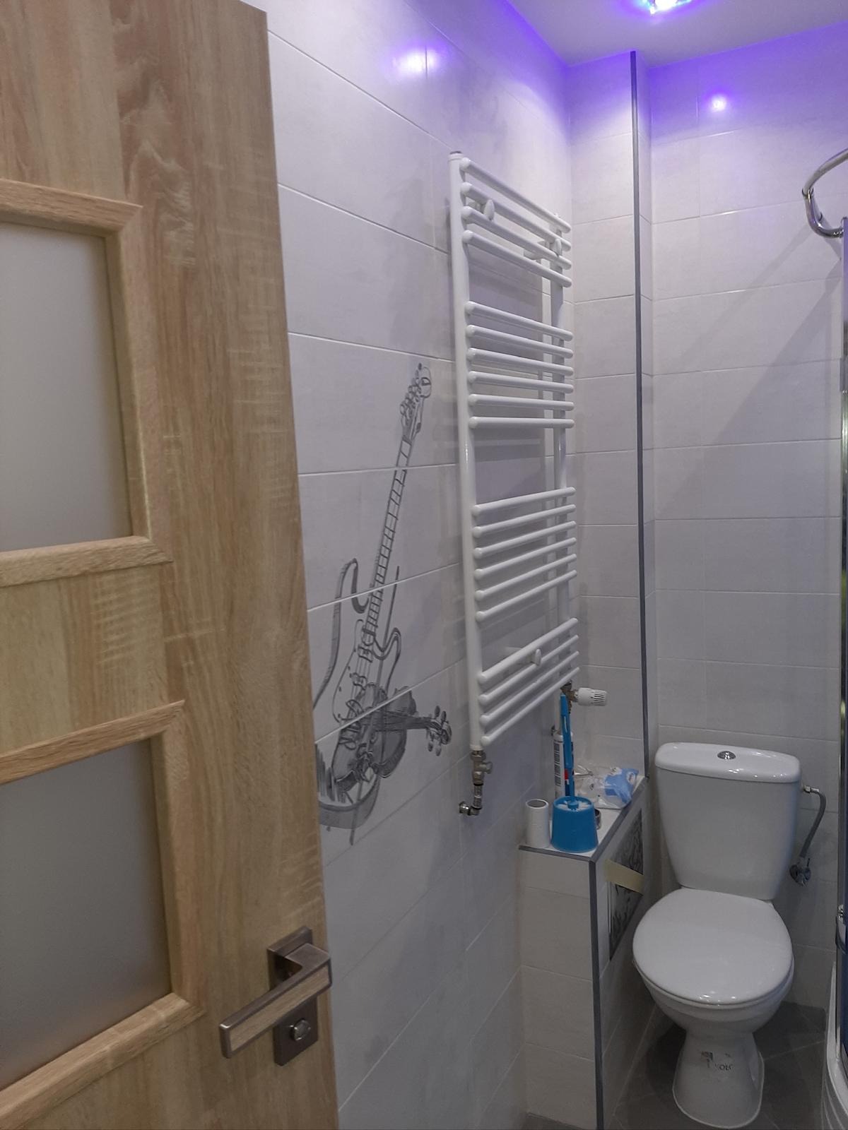 Wykończona łazienka z białą ceramiką, grzejnikiem drabinkowym i płytkami z motywem gitary. Widoczna drewniana ościeżnica drzwi i fioletowe oświetlenie sufitowe.