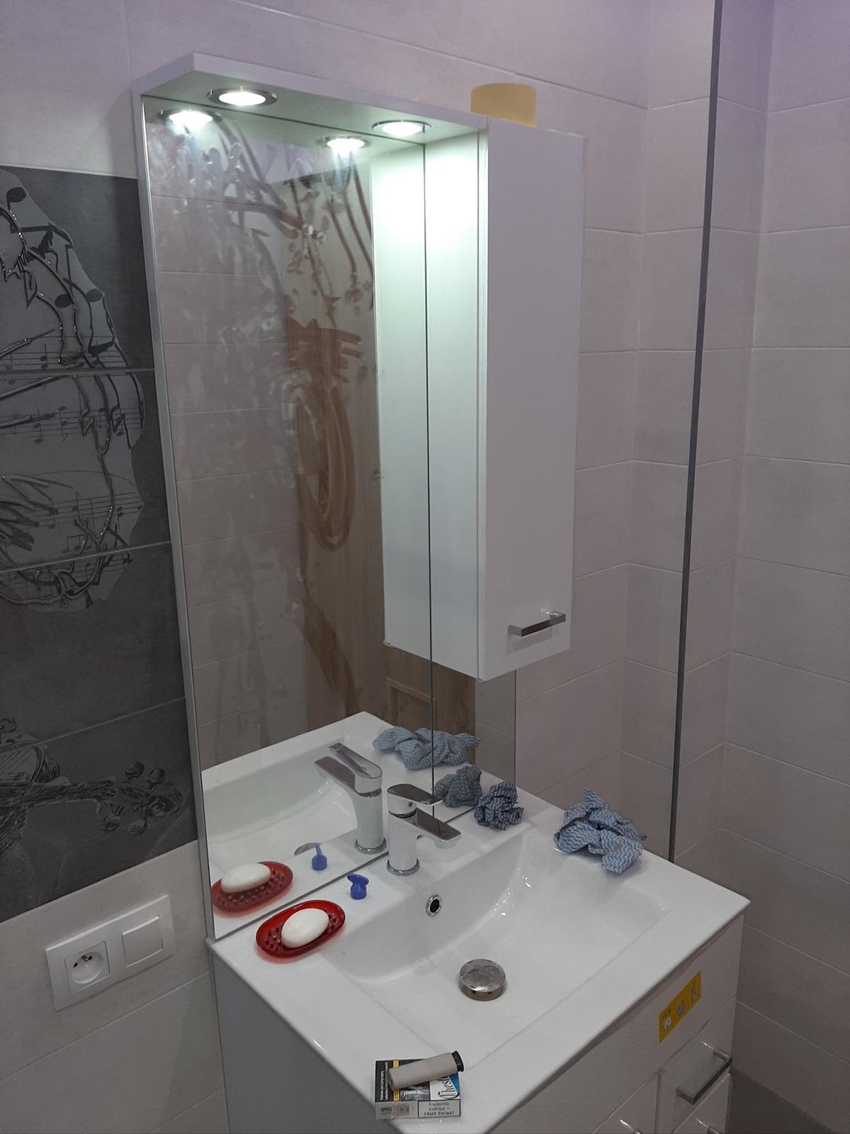 Wykończona łazienka z umywalką, lustrem z oświetleniem i szafką wiszącą. Na umywalce mydło w mydelniczce i ściereczki. Na ścianie płytki z motywem muzycznym.