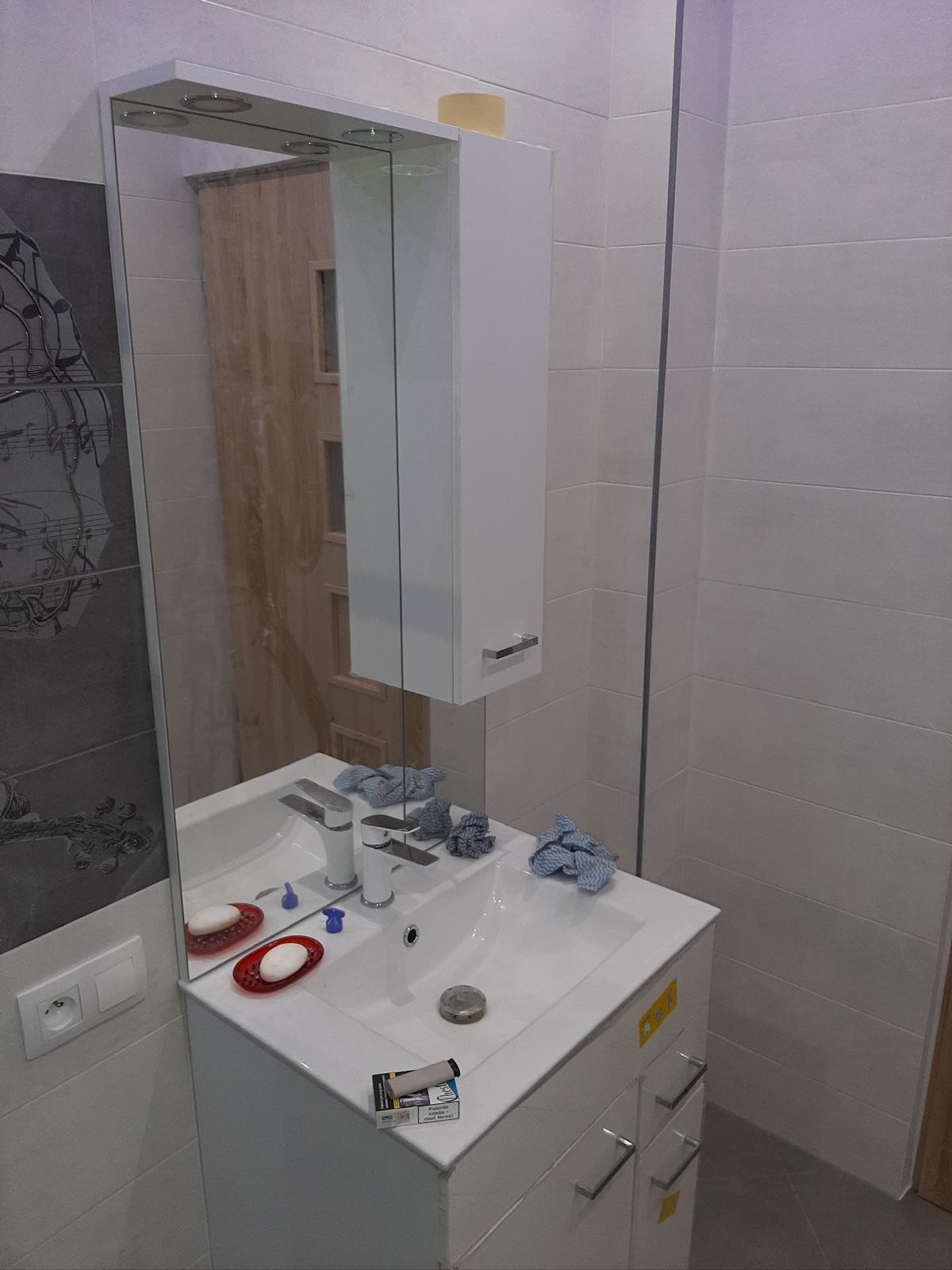 Biała umywalka z szafką i lustrem w łazience wykończonej jasnymi płytkami. Na blacie mydło i akcesoria, odbicie drzwi w lustrze.