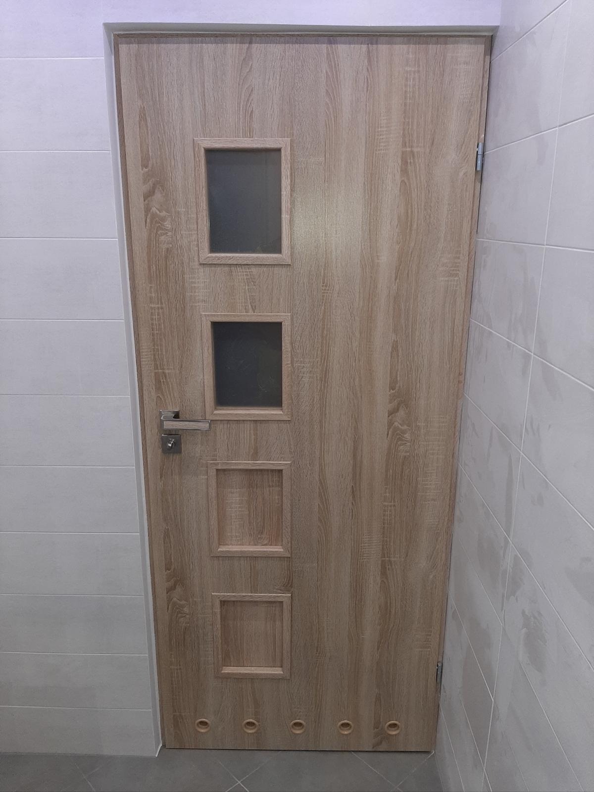 Zamknięte drzwi z okienkami w łazience z białymi płytkami. Drzwi w odcieniach beżu z imitacją drewna, trzy kwadratowe okienka, dwa z szybą, a jedno bez.