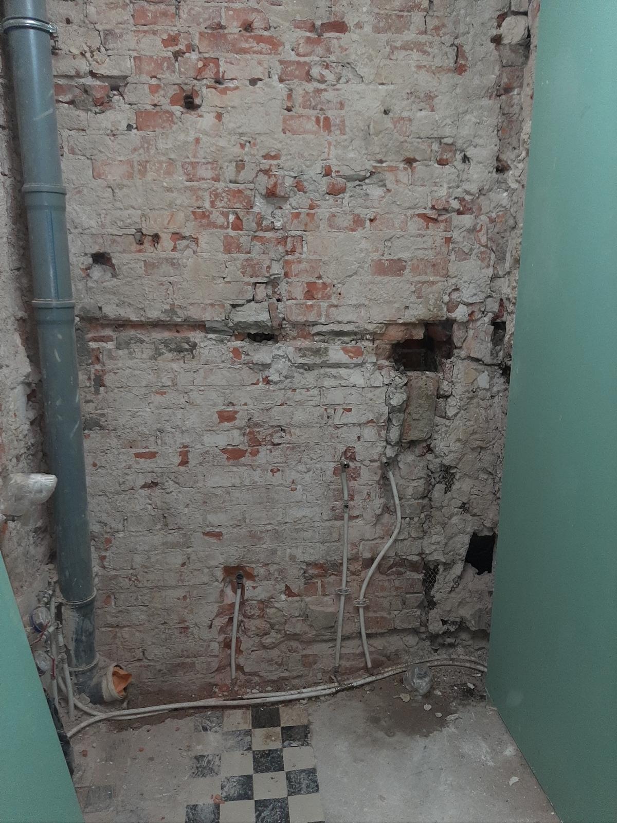 Odkryta ceglana ściana z instalacją hydrauliczną i fragmentem podłogi w szachownicę. Ściana w trakcie prac, widoczne uszkodzenia i pozostałości po tynku.