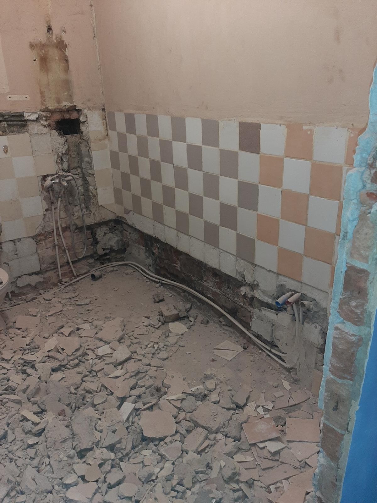 Demontaż płytek w łazience, widoczne rury i gruz. Fragment ściany z szachownicą z płytek. Przygotowanie do remontu, instalacja wodna.