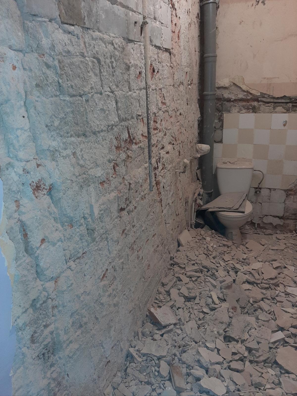 Łazienka w trakcie demontażu: odsłonięta cegła na ścianie, gruz na podłodze, widoczna toaleta z elementami starej instalacji. Przygotowanie do remontu.