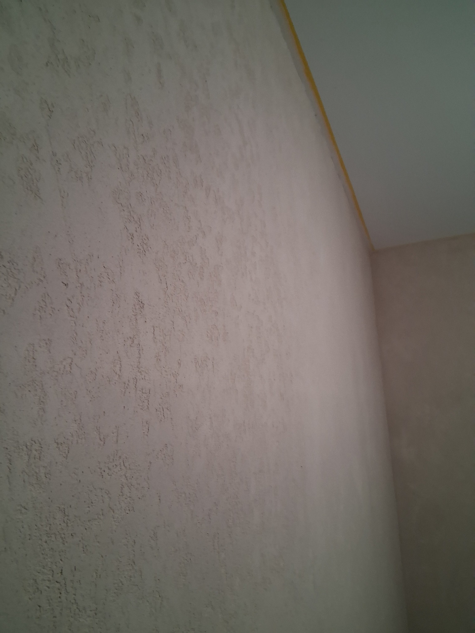 Ściana z fakturą w odcieniach beżu, widoczna taśma malarska przy suficie. Narożnik pokoju, sufit w jasnym kolorze. Tekstura ściany z bliska.