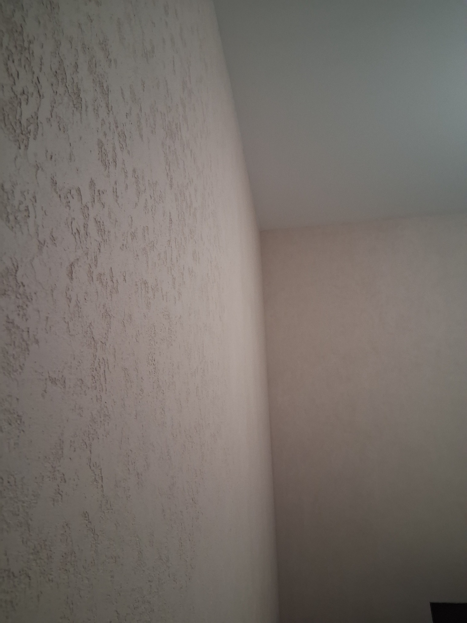 Ściana z dekoracyjną, beżową strukturą, widoczna pod kątem. Na ścianie widoczne nieregularne wzory i faktura. Górny róg kadru pokazuje fragment sufitu.
