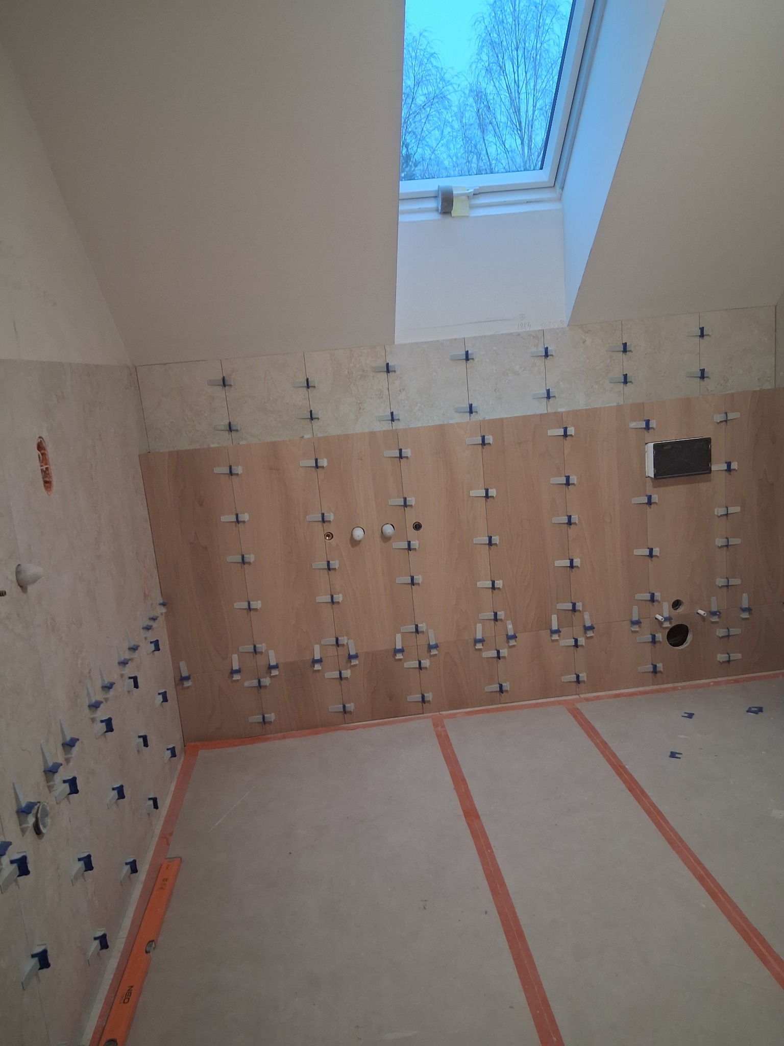 Ściana w trakcie układania płytek w łazience na poddaszu, widoczne krzyżyki dystansowe, otwory na instalacje i okno dachowe. Pomarańczowe linie na podłodze.
