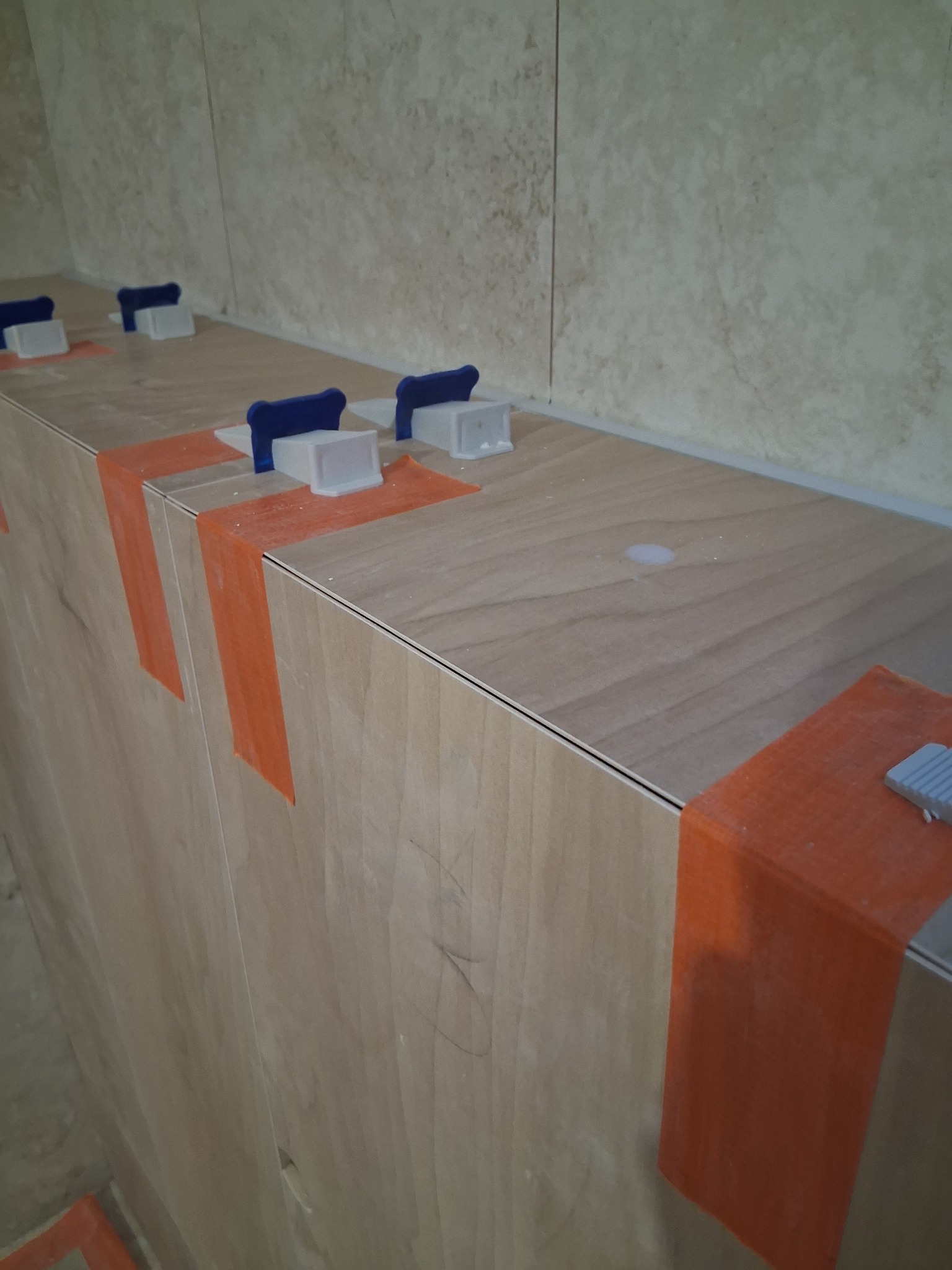 Układanie płytek na ścianie z użyciem systemu poziomowania: pomarańczowa taśma i niebieskie kliny. Widok z bliska na fragment ściany w trakcie prac wykończeniowych.