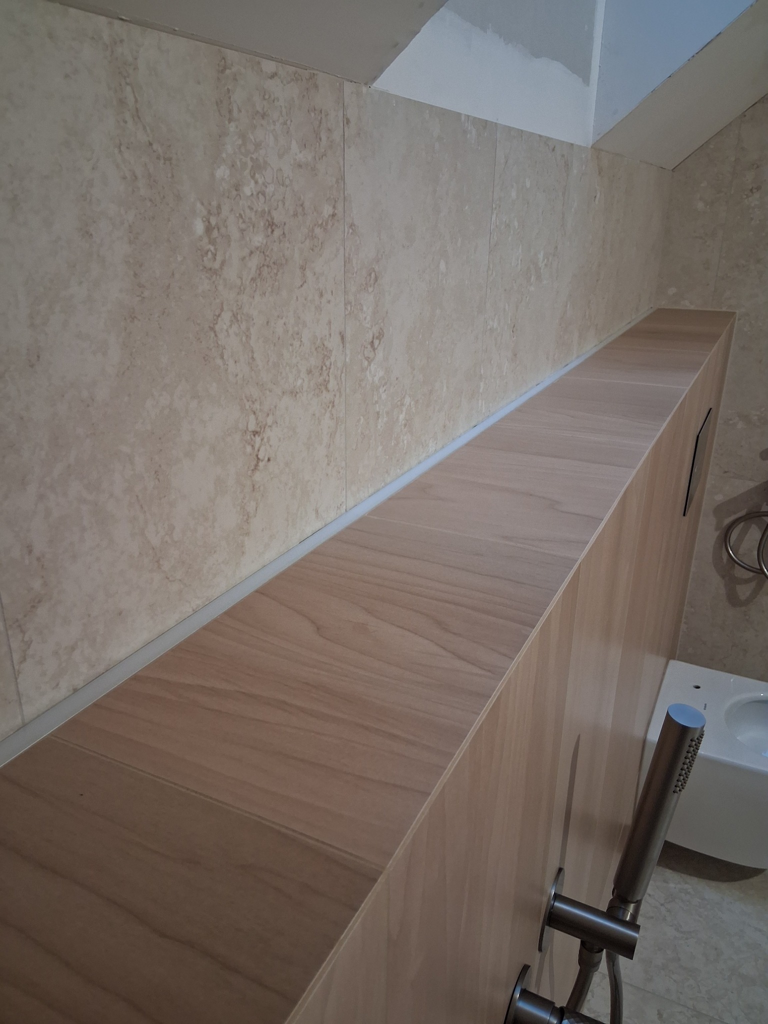 Wykończona łazienka z beżowymi płytkami imitującymi marmur i zabudową z drewnopodobnej płyty. Widoczna bateria prysznicowa i fragment toalety.