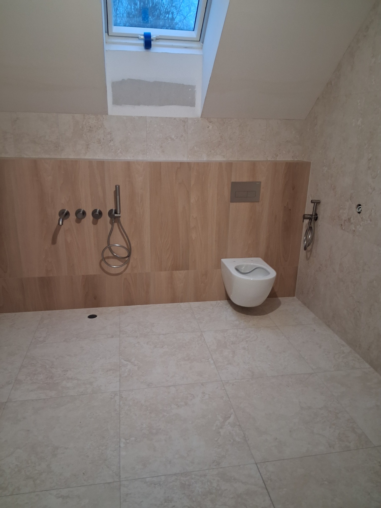 Nowoczesna łazienka z beżowymi płytkami, wiszącą toaletą i armaturą w kolorze stali. Okno dachowe wpuszcza naturalne światło. Minimalistyczny design.