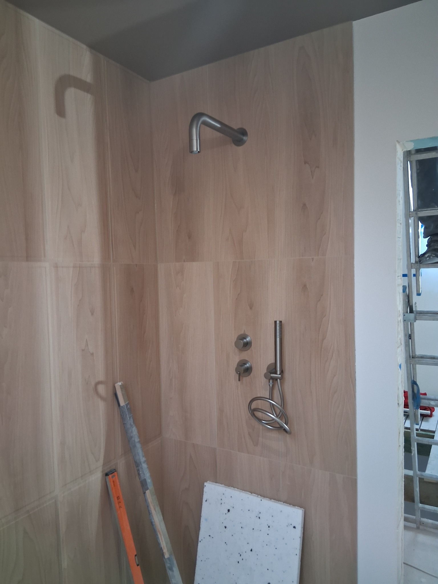 Wykończona kabina prysznicowa z drewnopodobnymi płytkami, widoczne narzędzia i drabina, sugerujące trwające prace wykończeniowe w łazience.