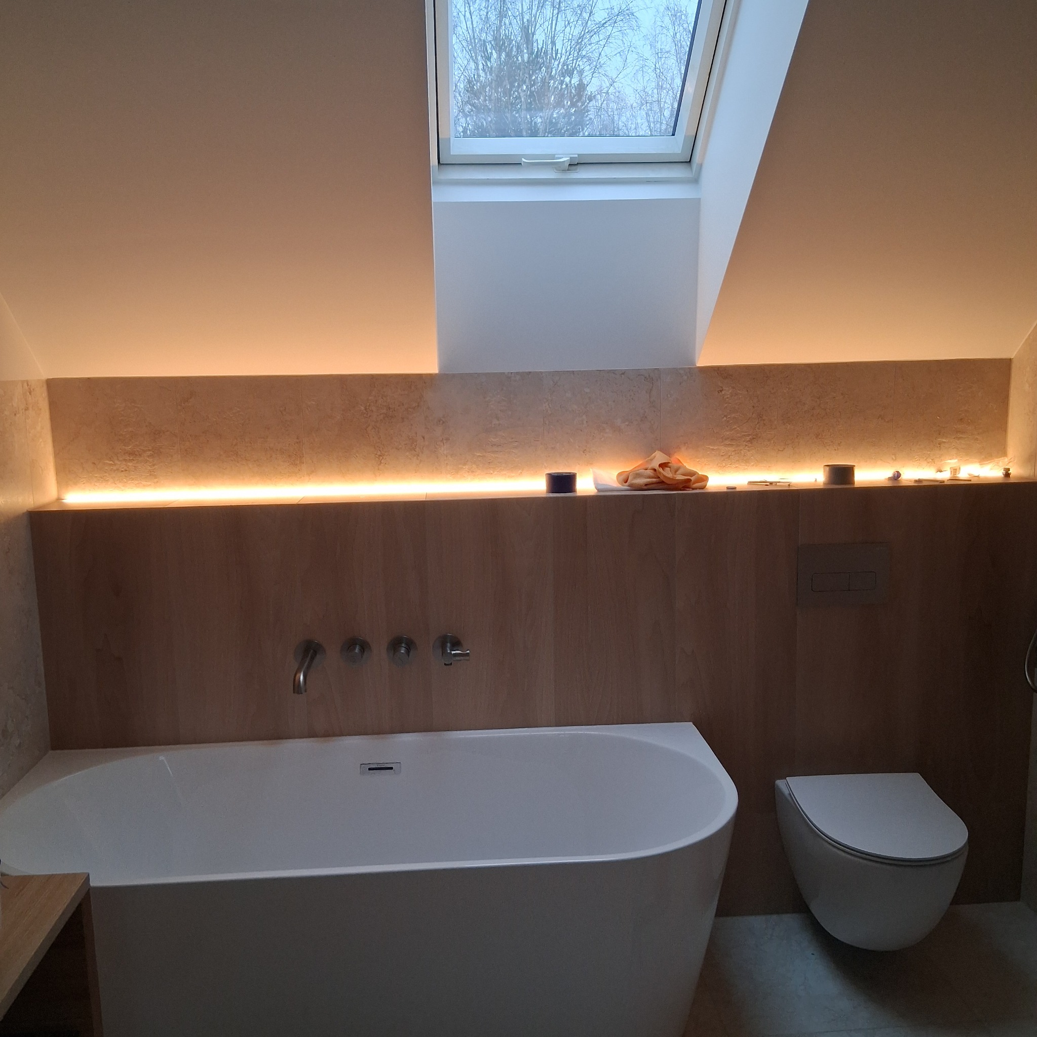 Nowoczesna łazienka na poddaszu z oknem dachowym, białą wanną i podwieszaną toaletą. Drewniane panele ścienne z subtelnym oświetleniem LED.