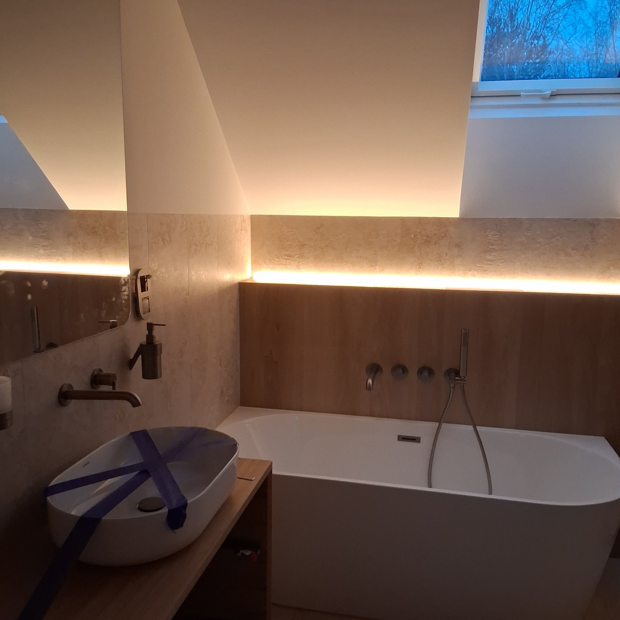 Nowoczesna łazienka na poddaszu z oknem dachowym. Umywalka owalna zabezpieczona taśmą, wanna prostokątna, beżowe płytki i oświetlenie LED.