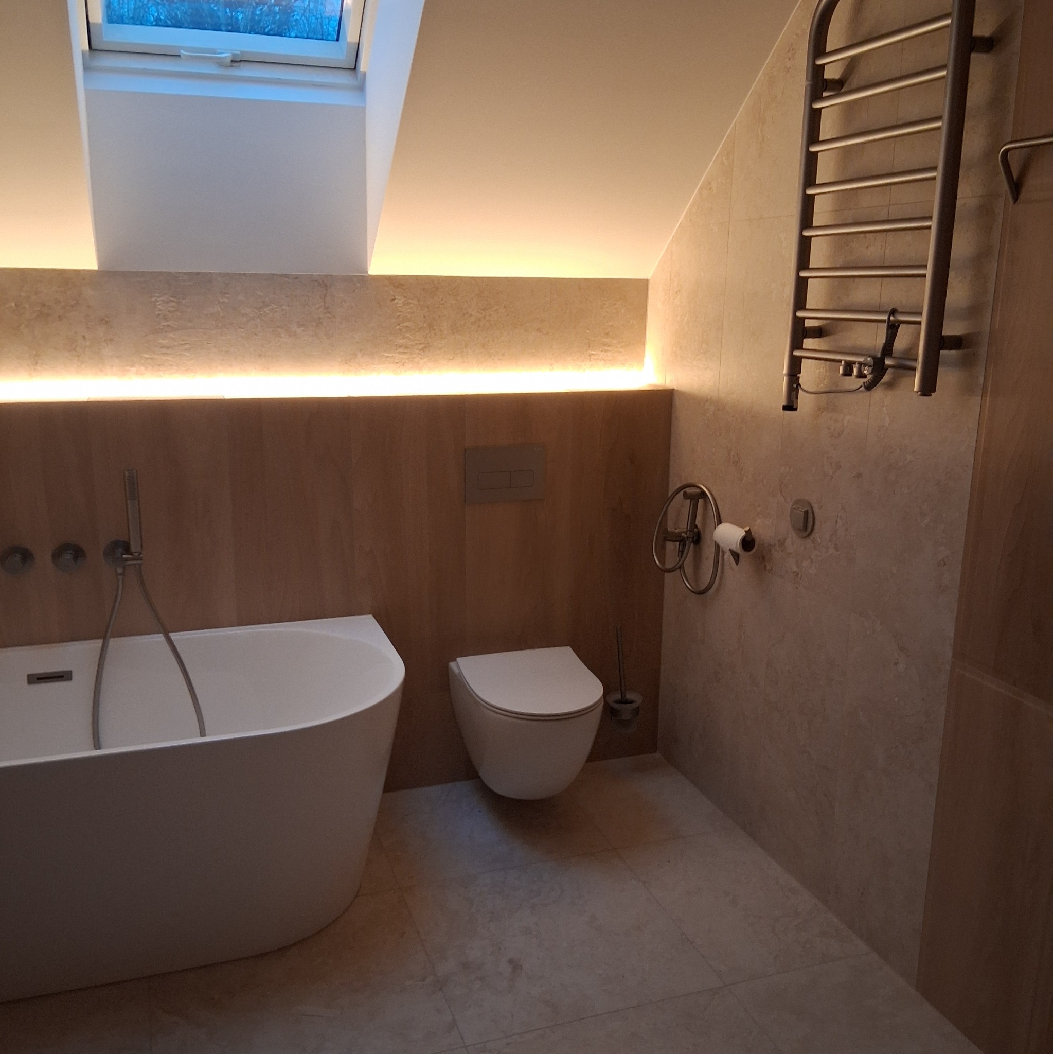 Nowoczesna łazienka w beżowych tonacjach z podświetleniem LED, owalną wanną i grzejnikiem drabinkowym. Widoczne okno dachowe i minimalistyczna armatura.