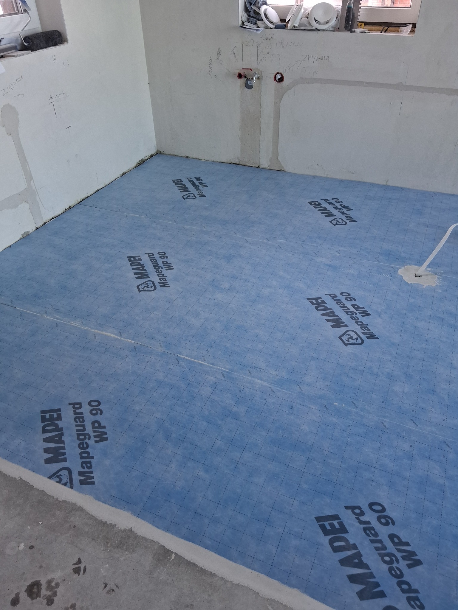 Niebieska hydroizolacja Mapeguard WP 90 na podłodze w surowej łazience przed położeniem płytek. Widoczne ślady po instalacjach na ścianach.