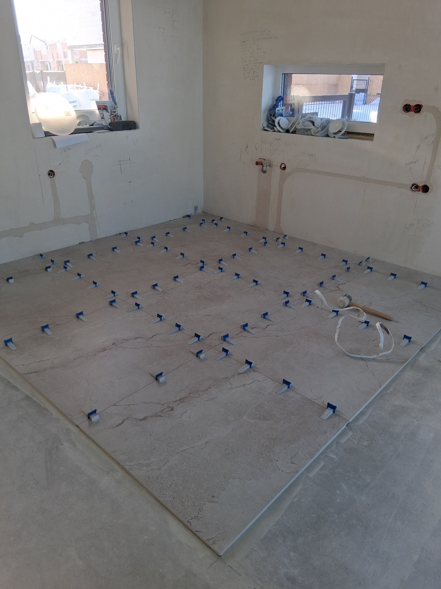 Układanie płytek podłogowych w jasnym odcieniu, z użyciem niebieskich krzyżyków dystansowych i gumowego młotka. Ściany z widocznymi instalacjami i notatkami.