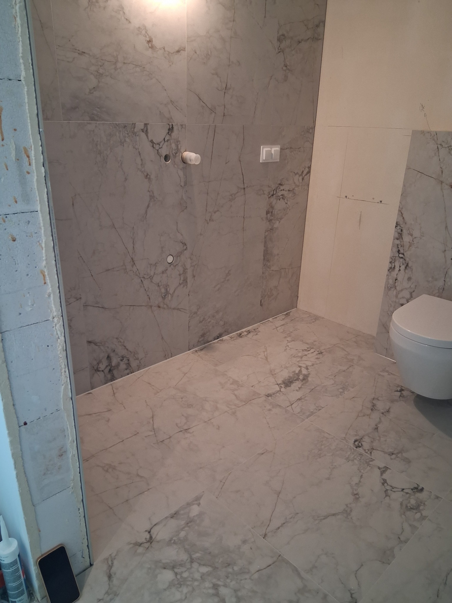 Łazienka w trakcie wykończenia: marmurowe płytki na ścianach i podłodze, widoczne instalacje, częściowo zamontowana toaleta. Prace glazurnicze w toku.
