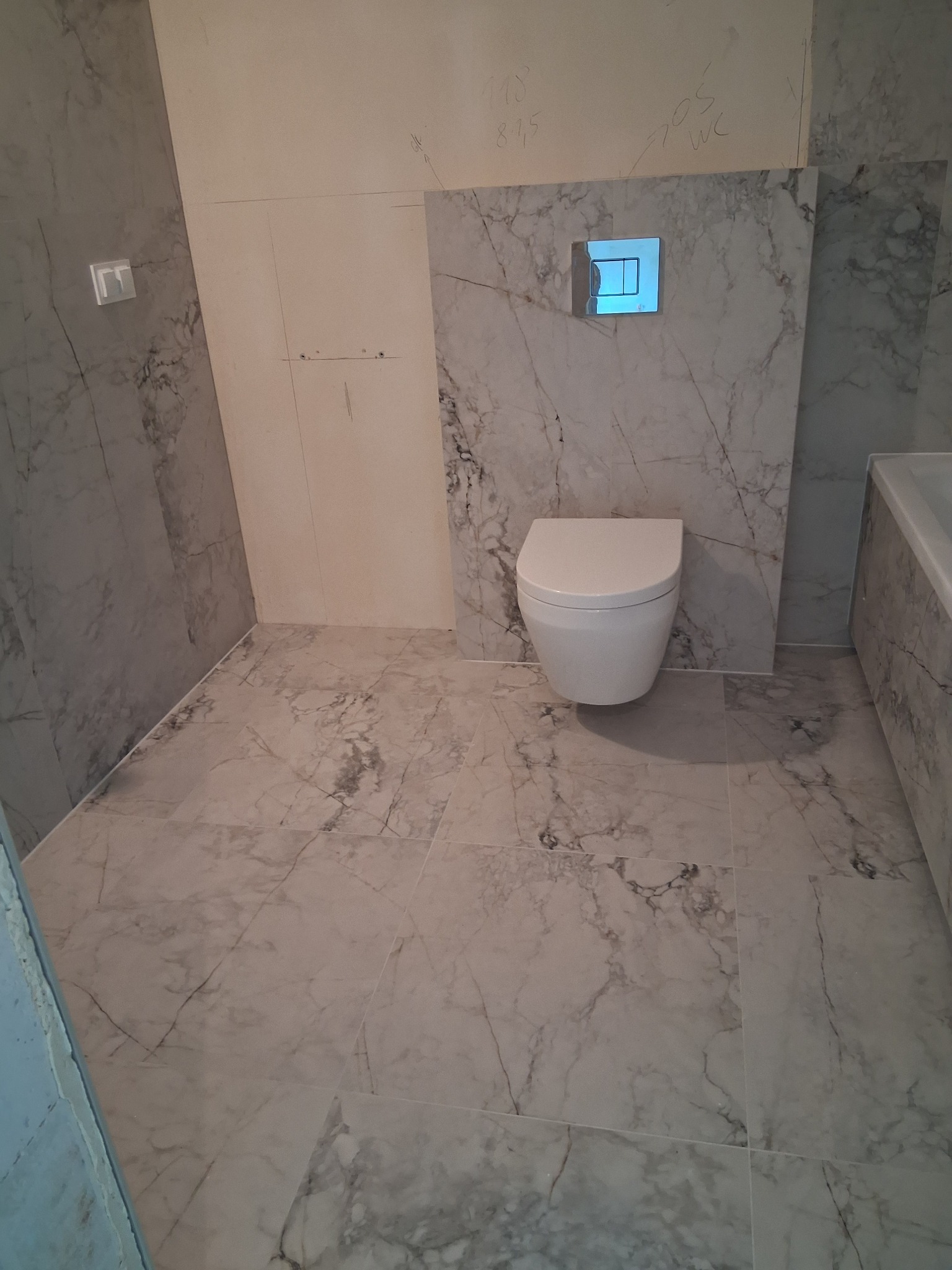 Wykończona łazienka z marmurowymi płytkami na ścianach i podłodze. Wisząca toaleta z chromowanym przyciskiem spłukującym. Widoczne ślady ołówka na ścianie.