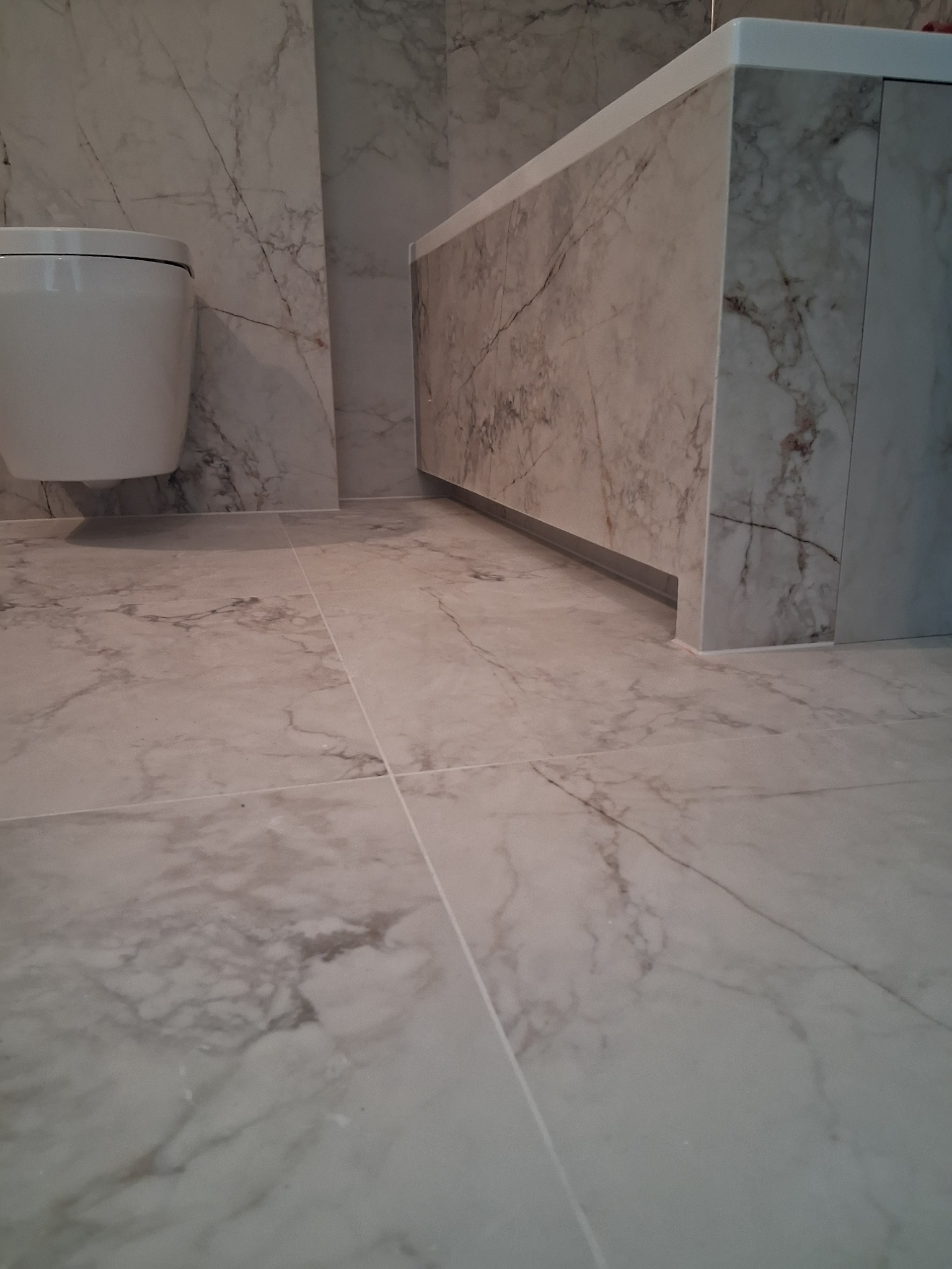 Wykończona łazienka z marmurowymi płytkami na ścianach i podłodze. Widoczna toaleta i zabudowa wanny, również wykończone marmurem. Minimalistyczny design, jasne wnętrze.