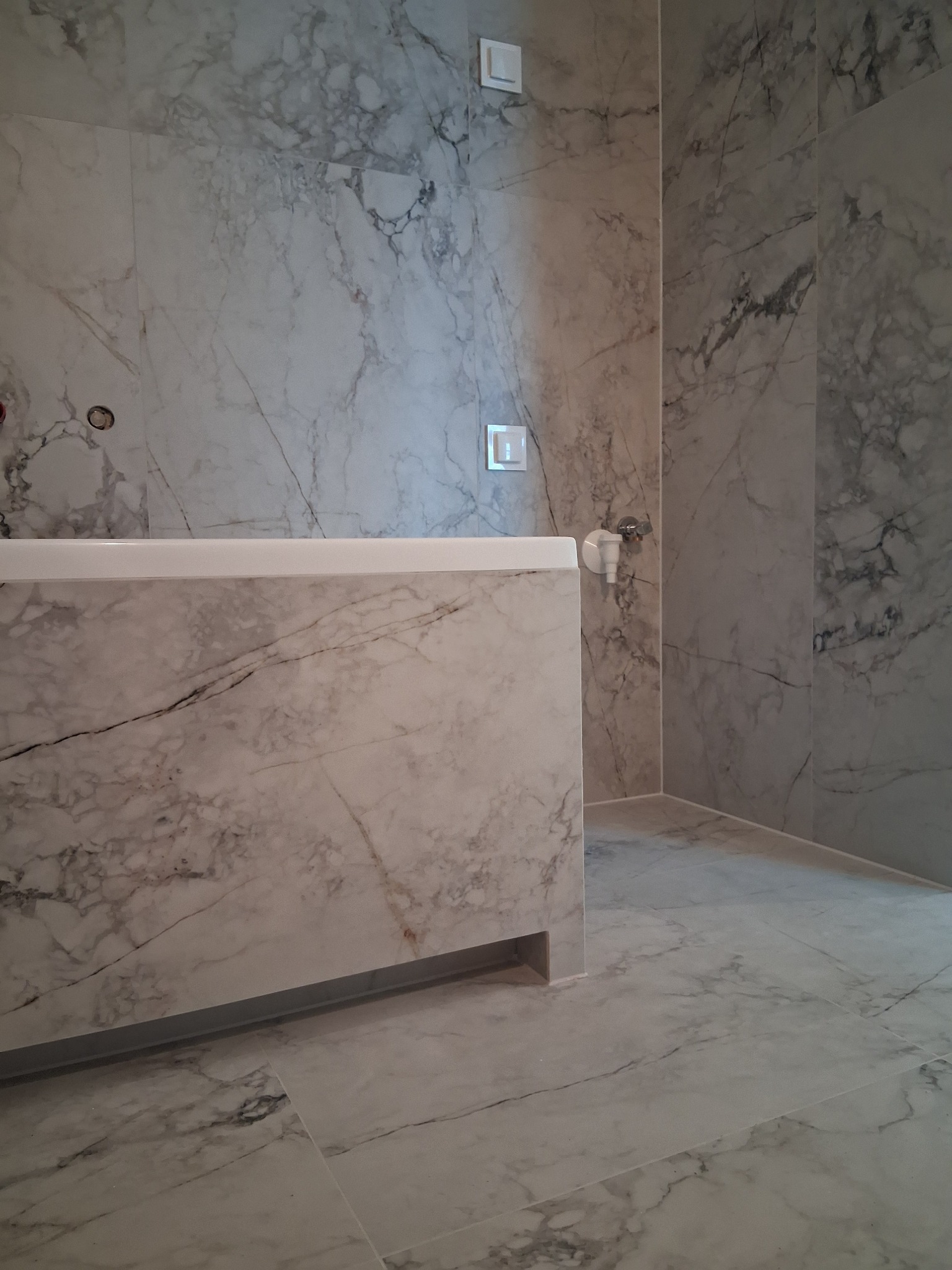 Wykończona łazienka z marmurowymi płytkami na ścianach i podłodze. Widoczna zabudowa wanny z tego samego materiału. Minimalistyczny design, jasne wnętrze.