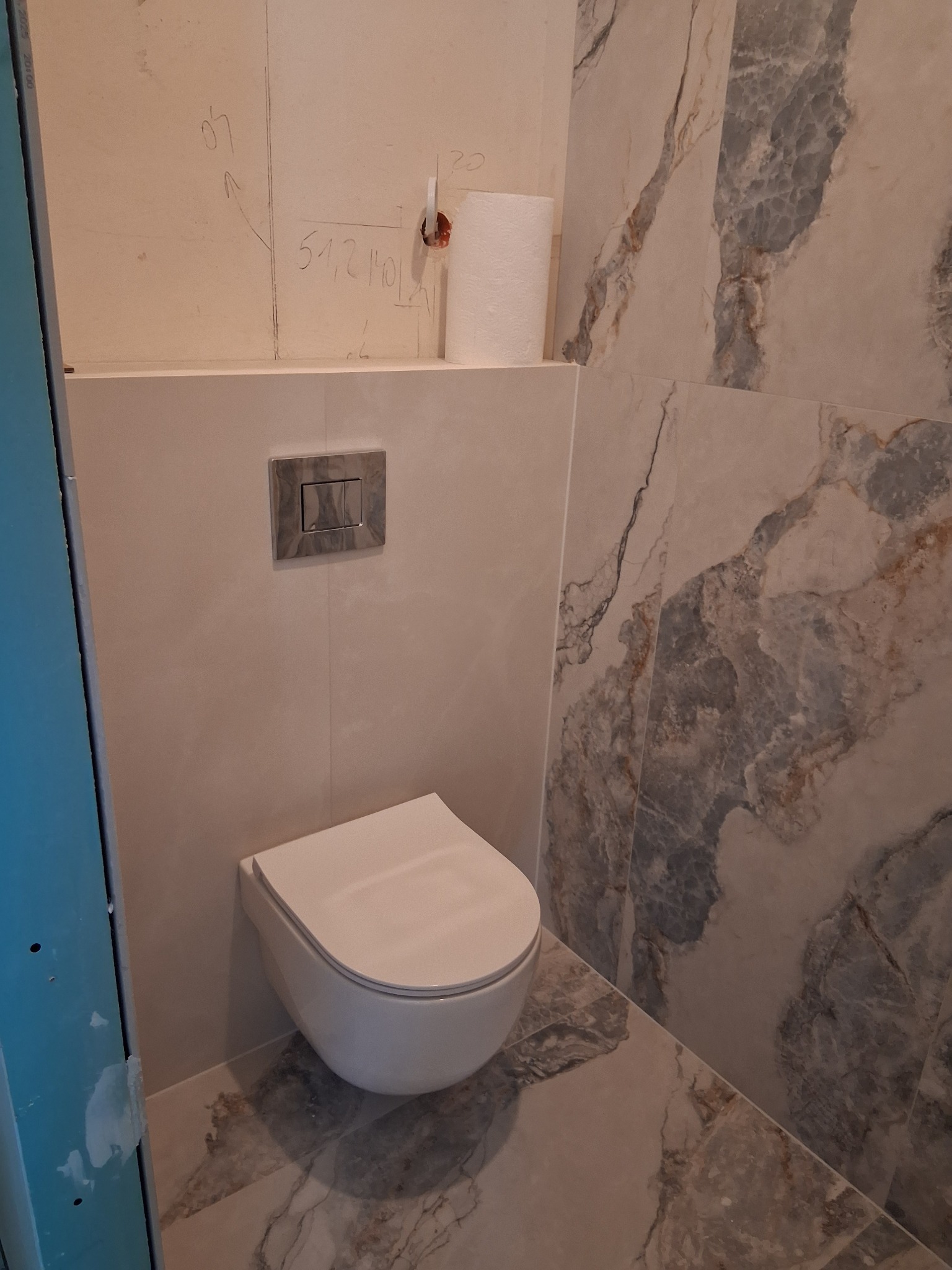 Wisząca toaleta w wykończonej łazience z marmurowymi płytkami na ścianach i podłodze. Widoczne oznaczenia na ścianie nad zabudową stelaża.