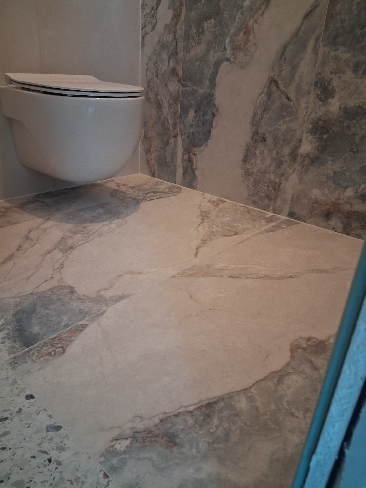 Wykończona łazienka z podwieszaną toaletą i marmurowymi płytkami na ścianach i podłodze. Widoczne detale fug i precyzyjne ułożenie płytek.