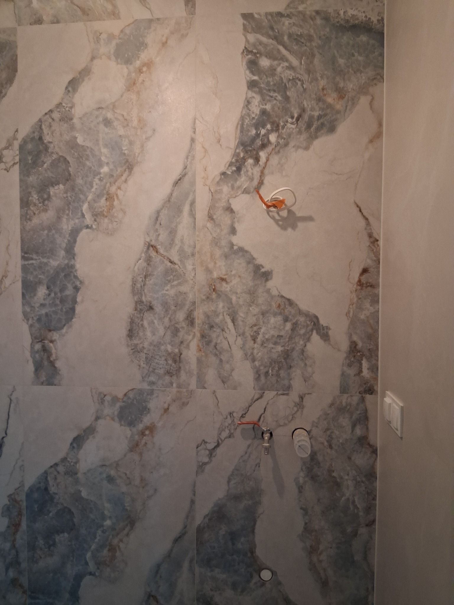 Ściana wyłożona marmurkowymi płytkami w odcieniach szarości i beżu, z widocznymi elementami instalacji wodnej i elektrycznej. Fragment ściany pomalowany na jednolity kolor.