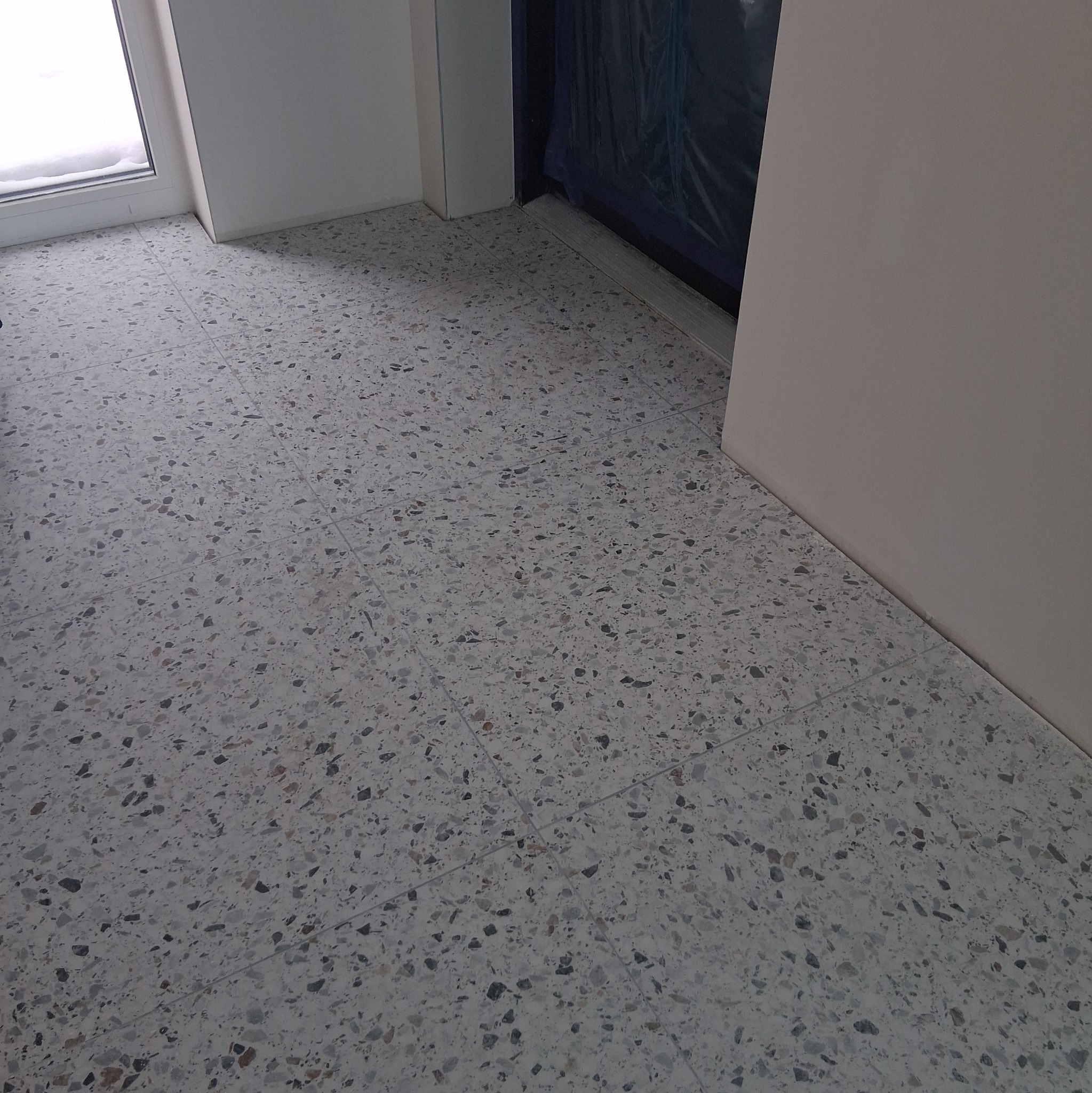 Szara, wzorzysta posadzka z terazzo w jasnym pomieszczeniu, widok z góry. Fragment okna i drzwi w tle. Minimalistyczny design podłogi.