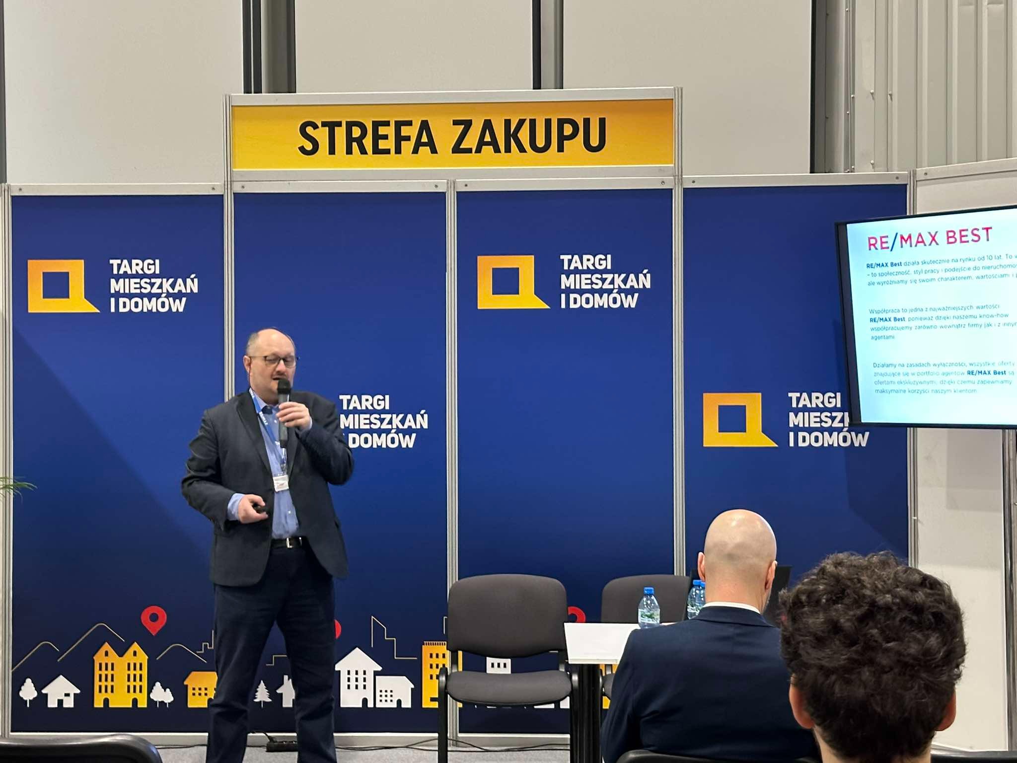 Prelegent z mikrofonem na tle stoiska Targów Mieszkań i Domów z napisem 'Strefa Zakupu'. Widoczny ekran z logo RE/MAX Best i uczestnicy.