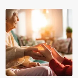 Dłoń opiekuna wspiera dłoń starszej kobiety w słonecznym pokoju, symbolizując troskę i bezpieczeństwo. Ciepłe barwy i bliskość rąk budują poczucie zaufania.