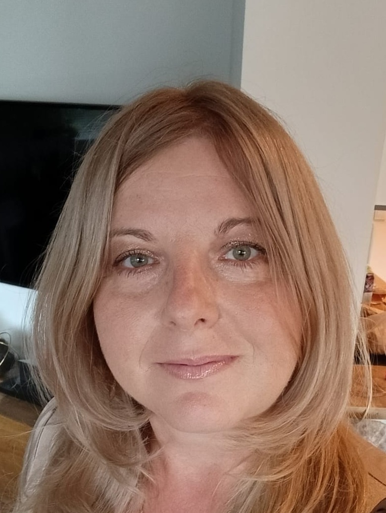 Portret uśmiechniętej kobiety z blond włosami, z delikatnym makijażem, patrzącej wprost w obiektyw. Widoczne tło wnętrza z telewizorem.