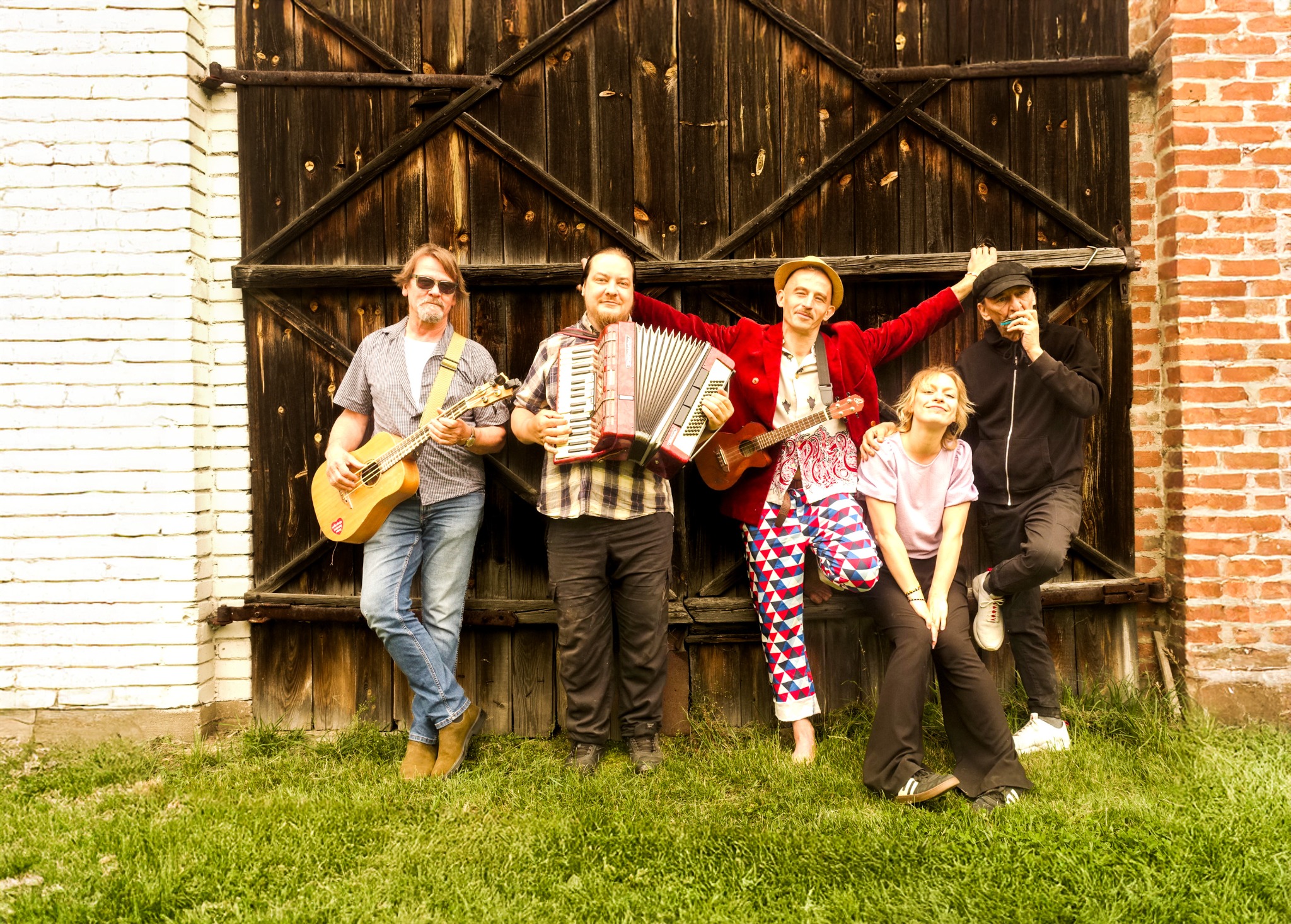 Zespół muzyczny w eklektycznych strojach pozuje z gitarami i akordeonem na tle drewnianych wrót i ceglanej ściany. Jeden z muzyków gra na harmonijce ustnej.