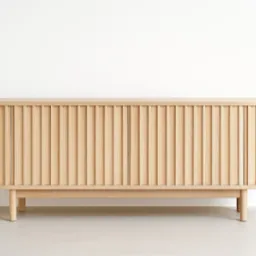 Drewniana komoda z ryflowanymi drzwiami, zaokrąglonymi krawędziami i minimalistycznymi nóżkami na tle białej ściany. Jasne drewno, prosta forma.