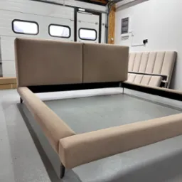 Beżowe, tapicerowane łóżko z wezgłowiem w garażu z białą bramą segmentową. Elementy łóżka w trakcie montażu, widoczne kartony i gaśnica.