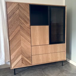 DANIEL KOPIEC DEKA FURNITURE - Nowoczesna, dębowa komoda z frezowanym wzorem w jodełkę na froncie, czarnymi akcentami i przeszkleniami, stojąca na smukłych, metalowych nóżkach.