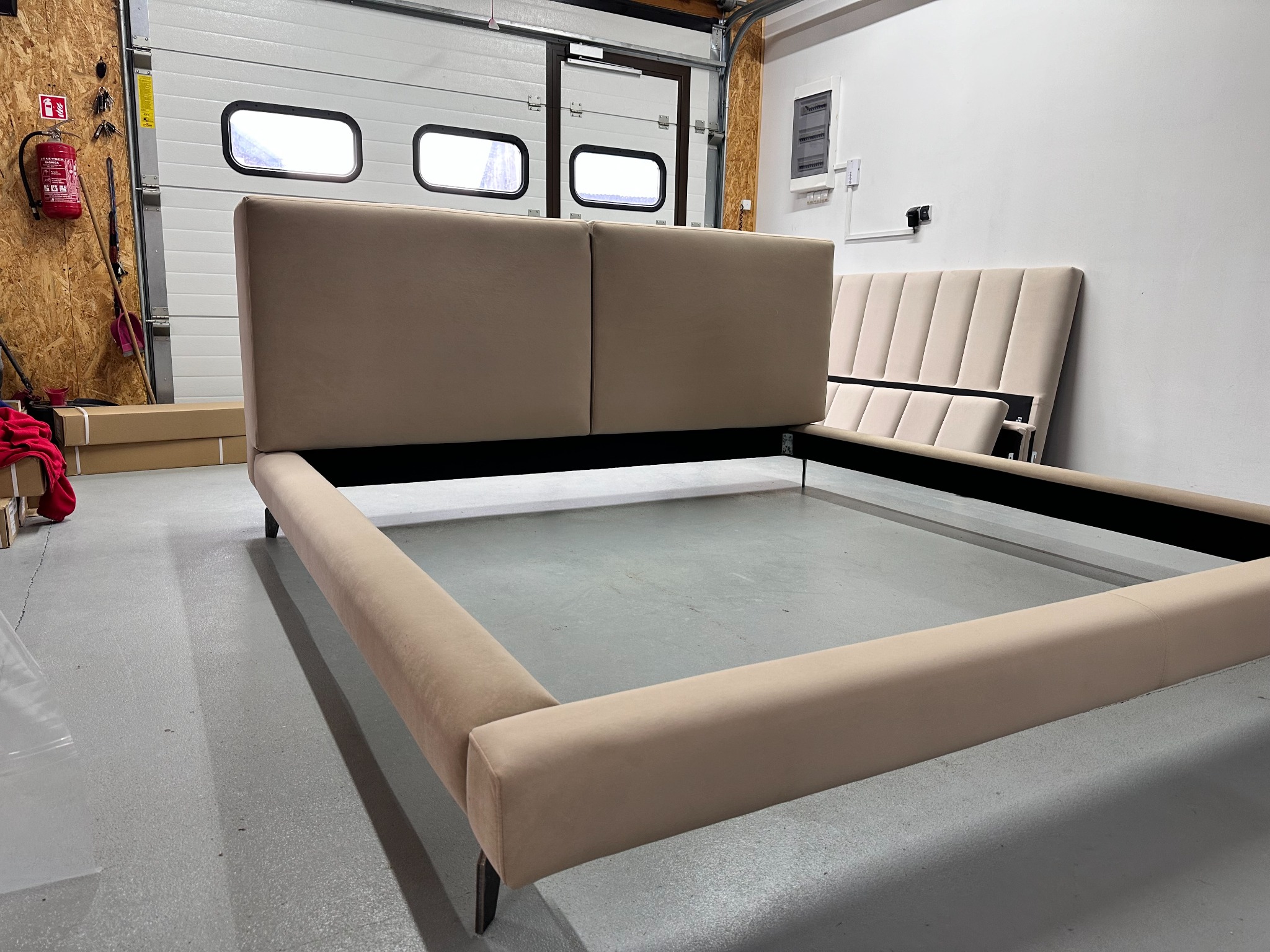 Beżowe, tapicerowane łóżko z wezgłowiem w garażu z białą bramą segmentową. Elementy łóżka w trakcie montażu, widoczne kartony i gaśnica.