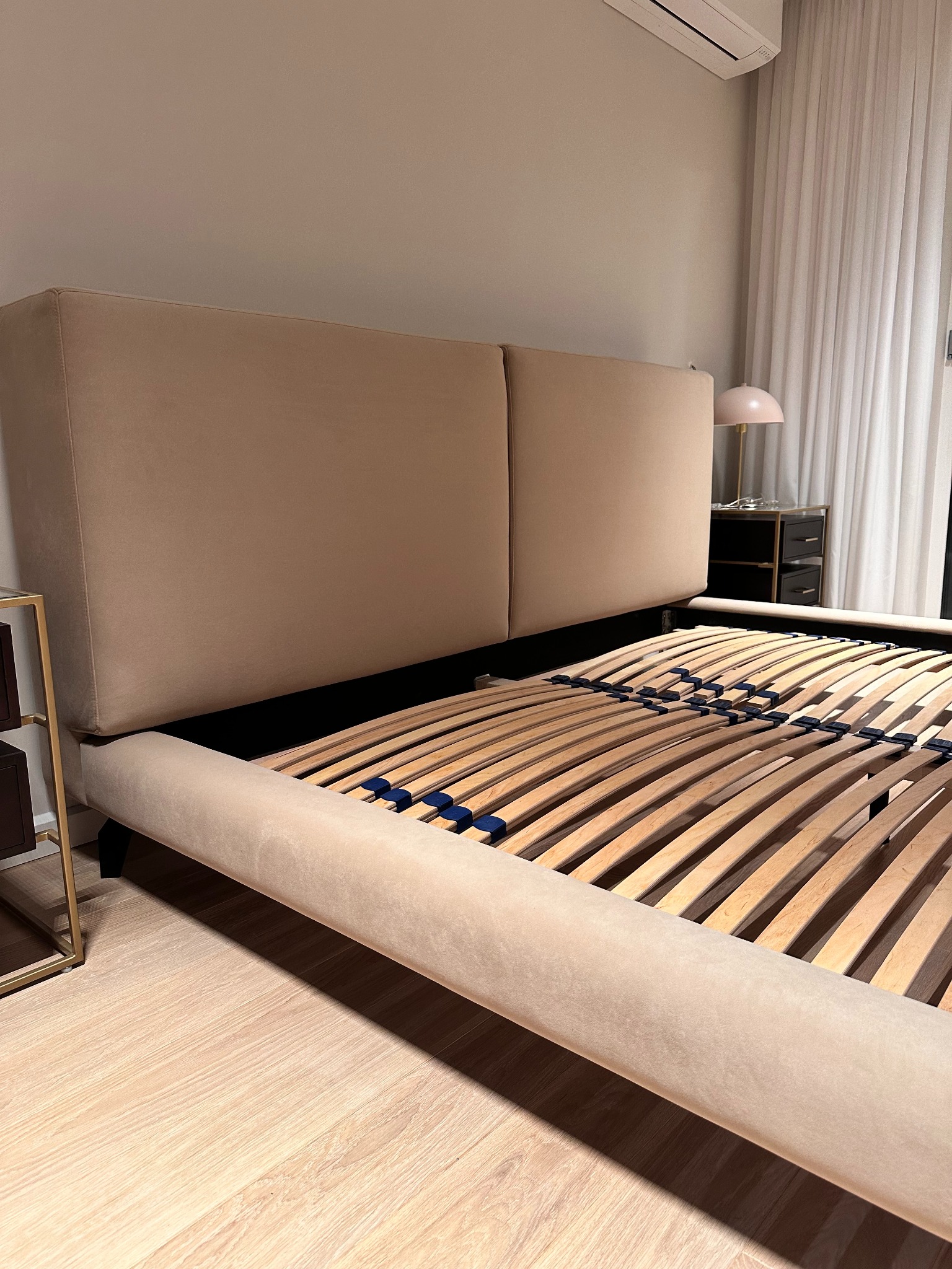 Beżowe łóżko z drewnianym stelażem i tapicerowanym zagłówkiem, widoczne z boku, z szafką nocną i lampką w tle.