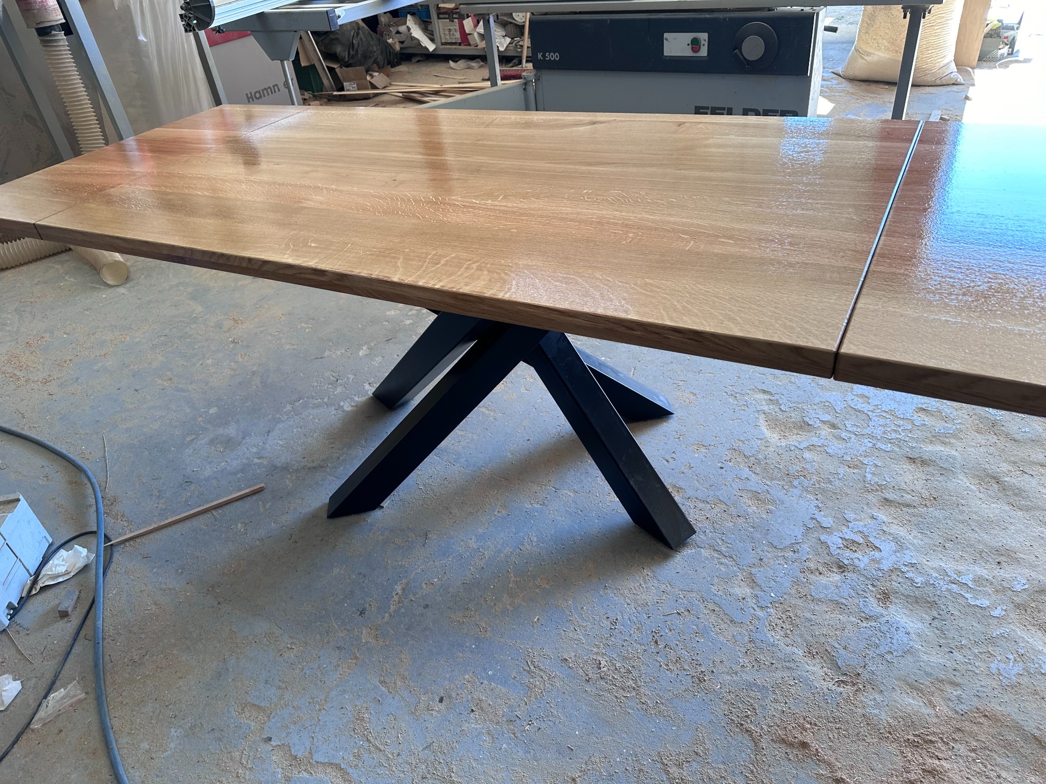 Rozkładany stół z blatem z jasnego drewna i czarnymi, geometrycznymi nogami, w tle warsztat stolarski z maszynami i narzędziami. Widoczny połysk lakieru na blacie.