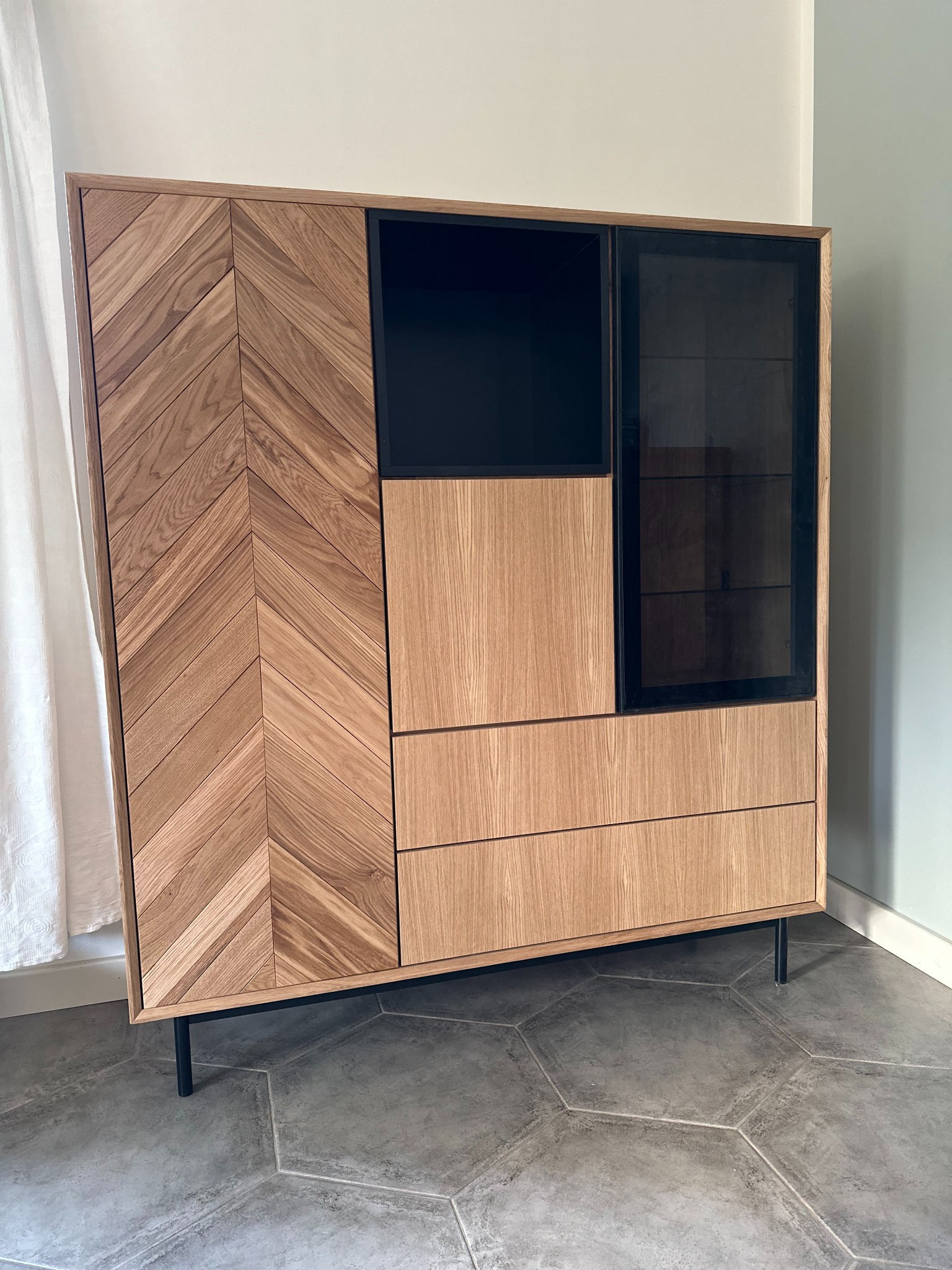 Nowoczesna, dębowa komoda z frezowanym wzorem w jodełkę na froncie, czarnymi akcentami i przeszkleniami, stojąca na smukłych, metalowych nóżkach.