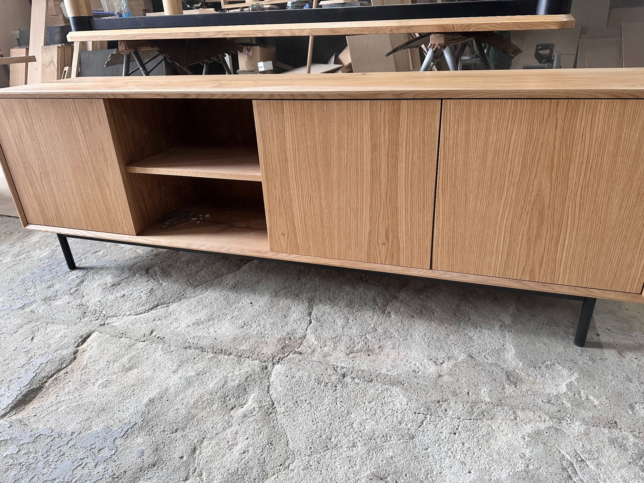 Drewniana komoda z przesuwnymi drzwiami i półkami, na metalowych nóżkach, w warsztacie. Wykończenie w naturalnym odcieniu drewna dębowego.