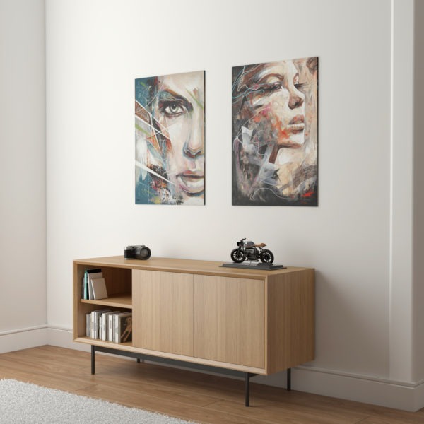 Jasna komoda z drewna na czarnych nóżkach, z przesuwanymi drzwiami. Na niej aparat, model motocykla. Nad nią obrazy z abstrakcyjnymi portretami.