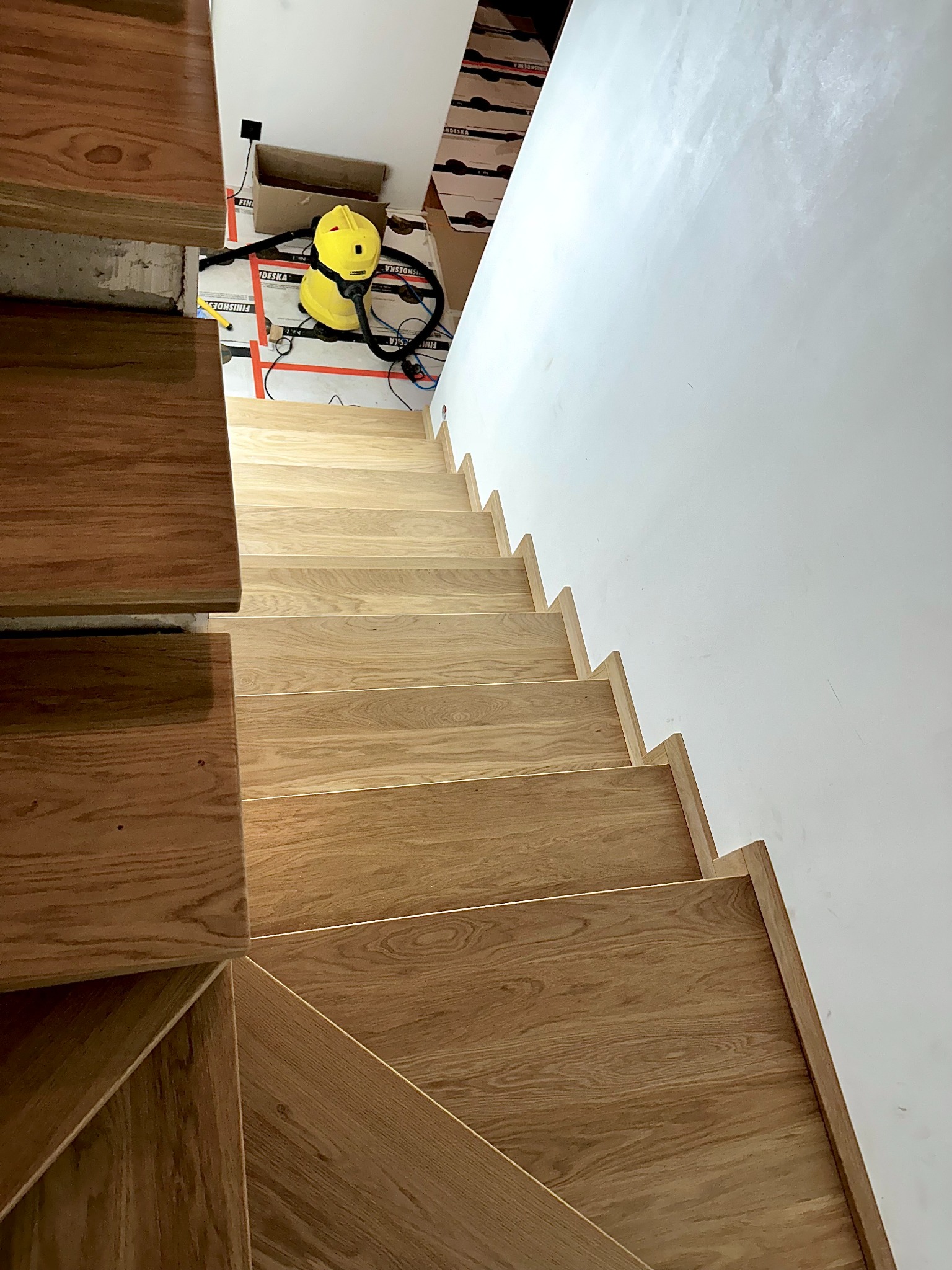 Jasne, drewniane schody z widocznym usłojeniem, prowadzące w górę wzdłuż białej ściany. W tle widoczny żółty odkurzacz i kartony, co sugeruje etap wykończeniowy.