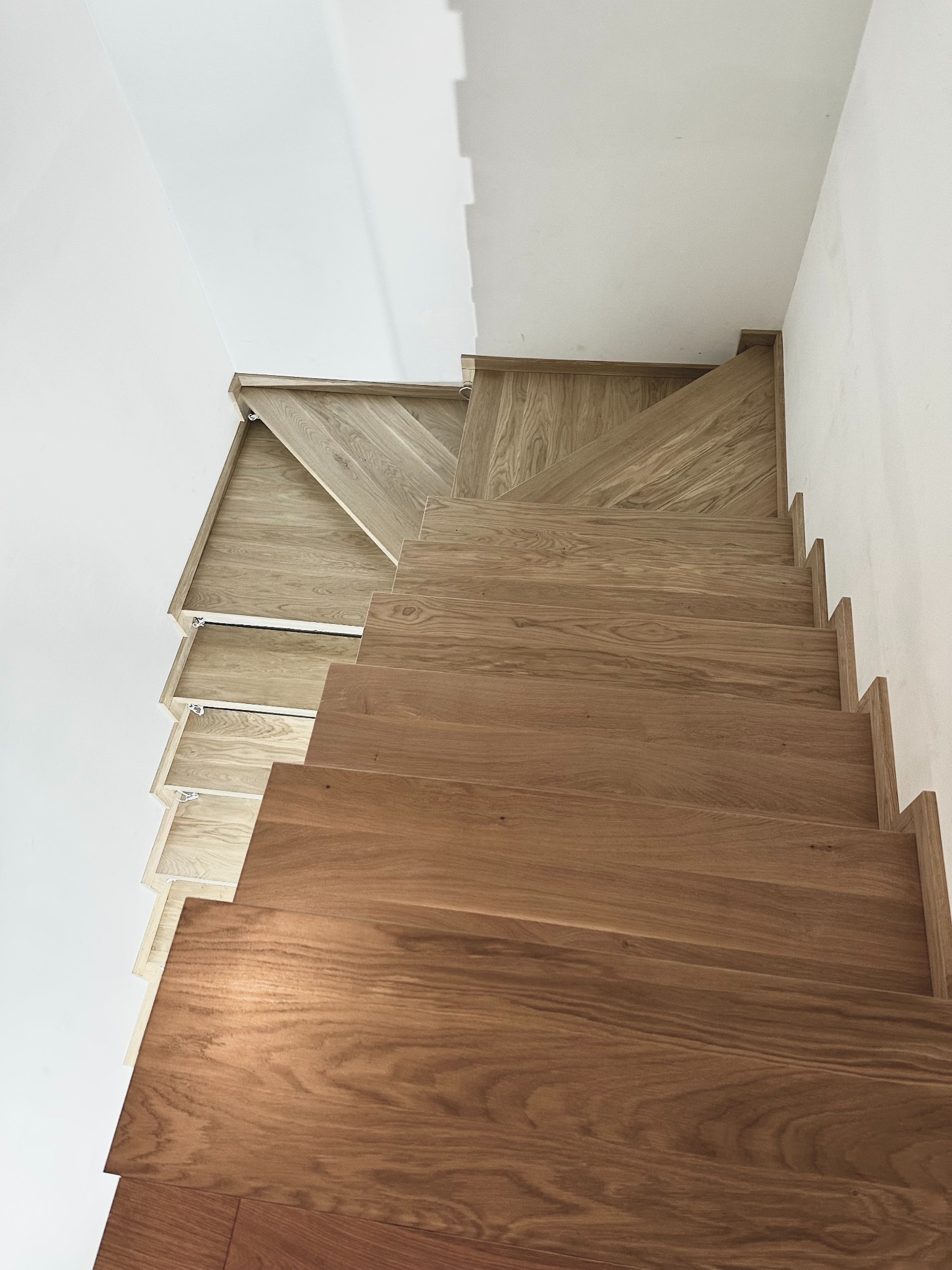 Drewniane schody z jasnego drewna, widok z góry, z białymi ścianami. Stopnie o zróżnicowanej kolorystyce drewna, od jasnego do ciemnego brązu. Minimalistyczny design.