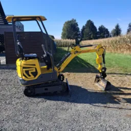 Żółta minikoparka Wacker Neuson na żwirowym podłożu, z łyżką skierowaną w dół, w tle pole kukurydzy i budynek. Słoneczny dzień.