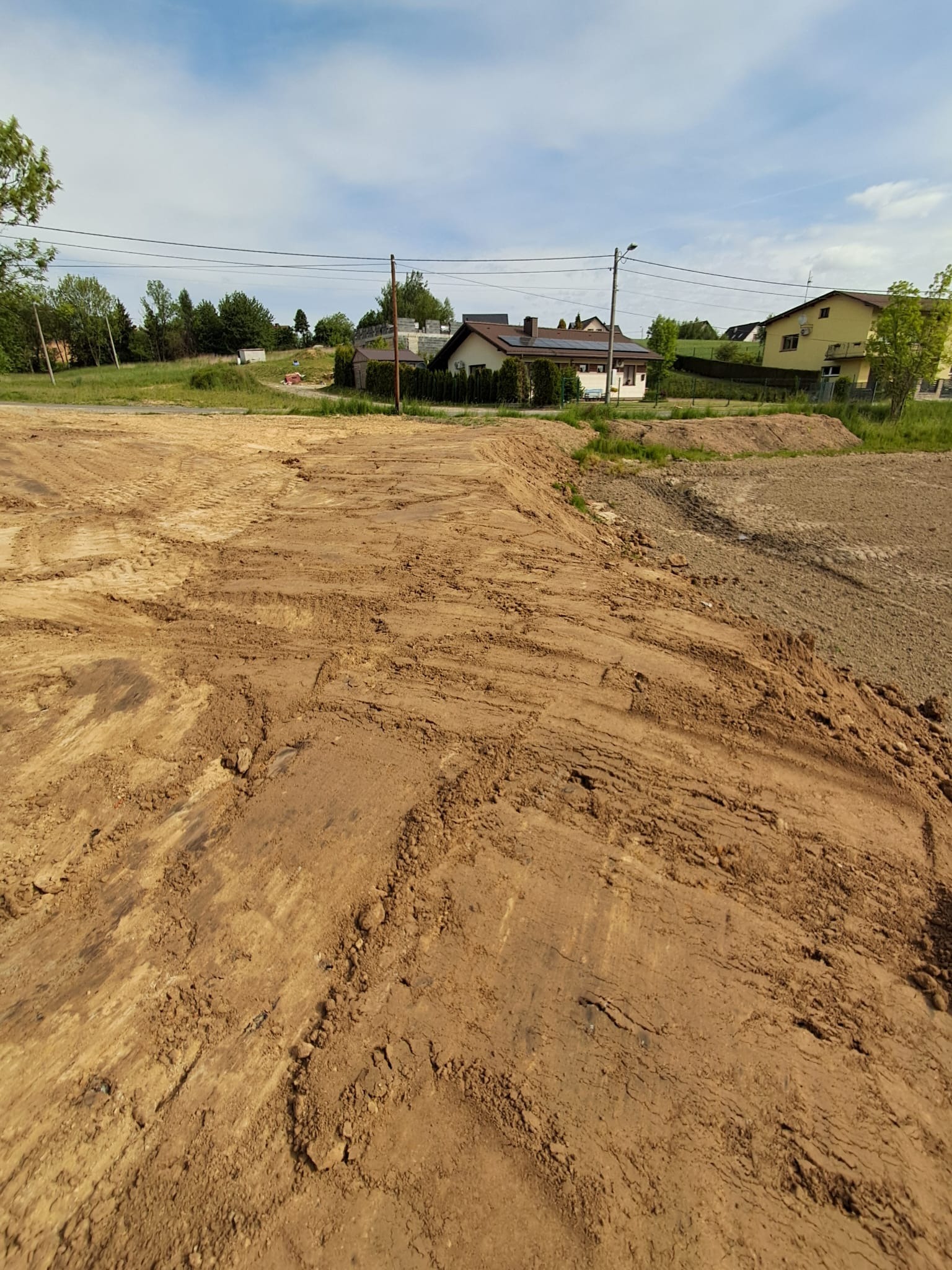 Nierówny, brązowy teren po niwelacji z widocznymi śladami kół, w tle domy jednorodzinne i zieleń pod błękitnym niebem. Krajobraz po robotach ziemnych.