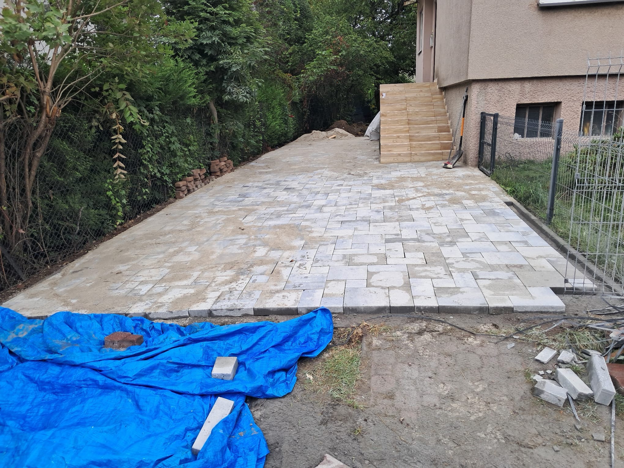 Układanie kostki brukowej na posesji w Rudzicy, widok na częściowo ułożony chodnik z szarych kostek, przygotowane podłoże i narzędzia. Prace w toku.