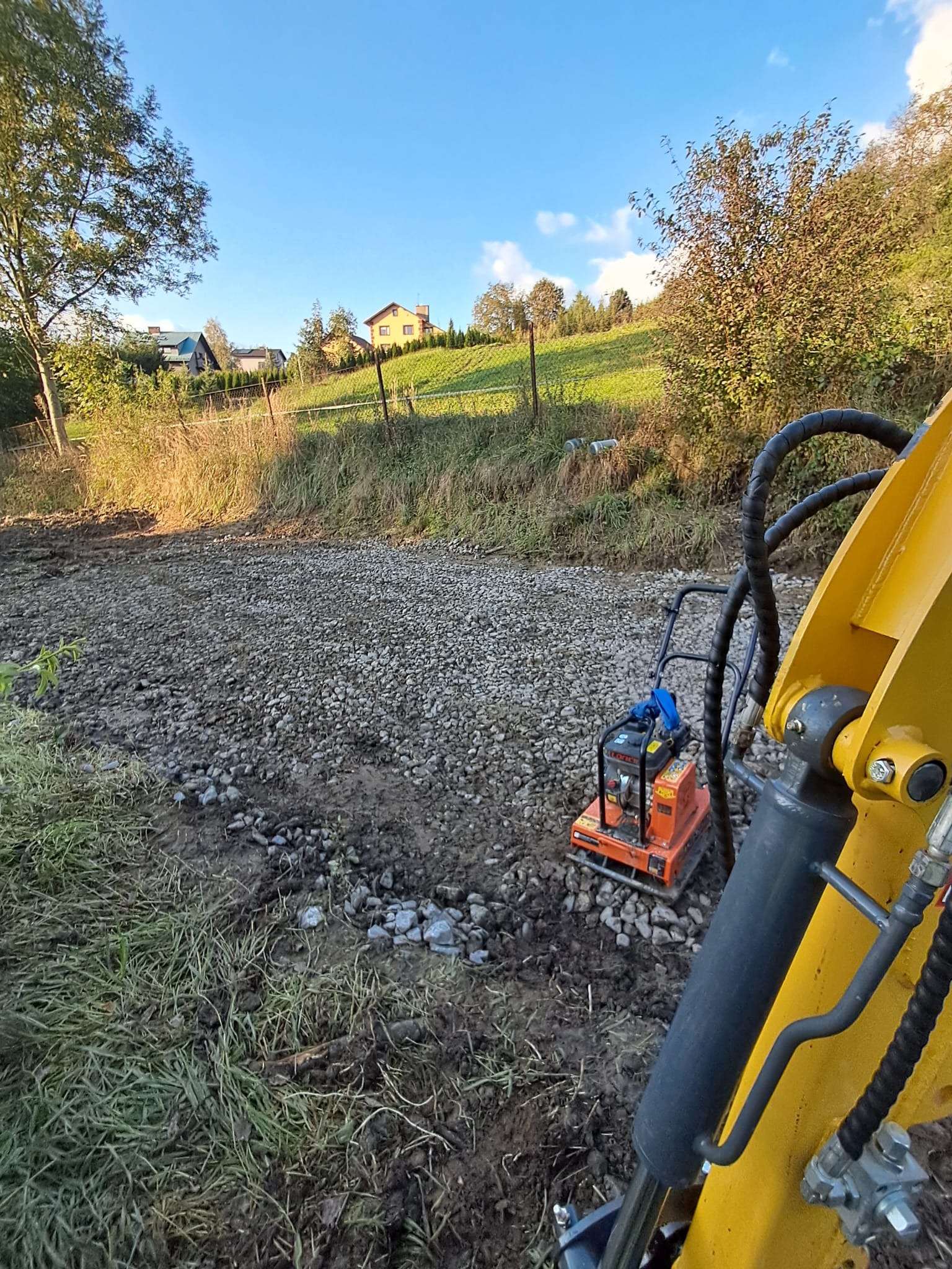 Pomarańczowa zagęszczarka spalinowa na żwirowej drodze polnej w trakcie utwardzania. W tle trawa, drzewa i zabudowania pod błękitnym niebem.