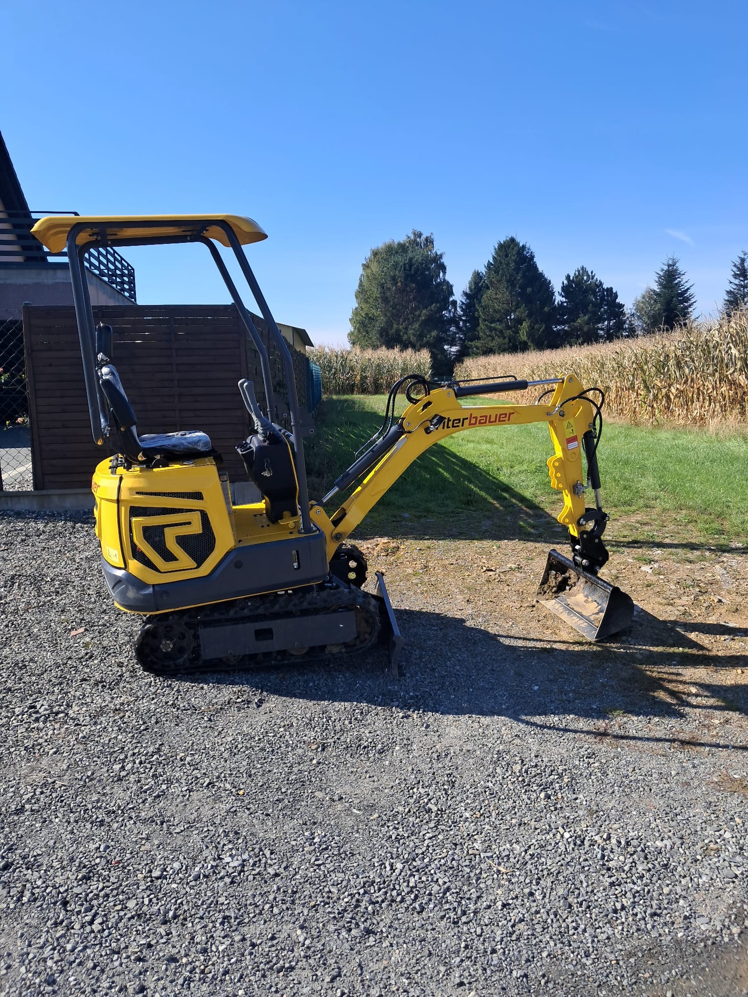 Żółta minikoparka Wacker Neuson na żwirowym podłożu, z łyżką skierowaną w dół, w tle pole kukurydzy i budynek. Słoneczny dzień.