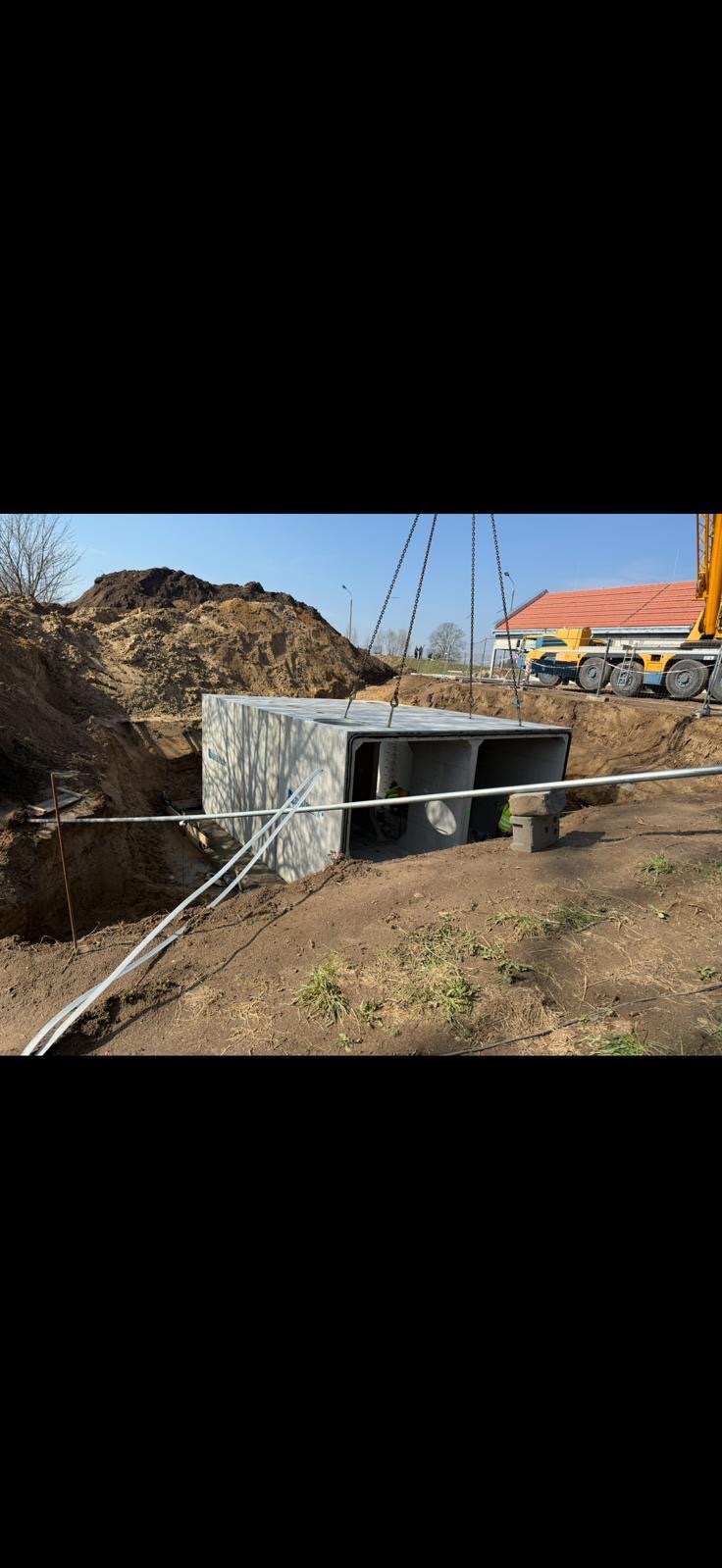 Montaż betonowego przepustu skrzynkowego w wykopie za pomocą dźwigu. Widoczne łańcuchy, wykop, hałda ziemi i fragment zabudowy w tle. Słoneczny dzień.