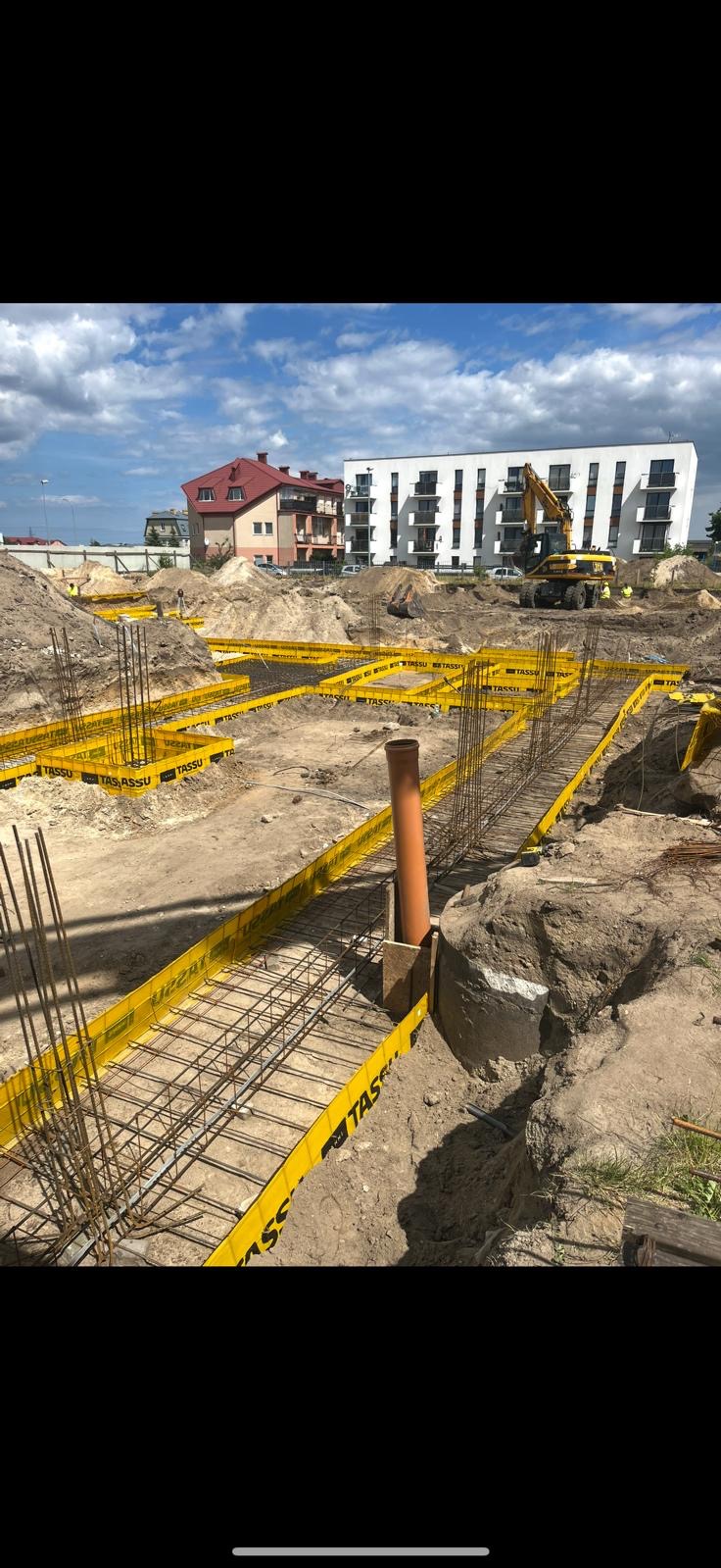 Zbrojenie fundamentów z żółtymi szalunkami TASSU na placu budowy, w tle budynki mieszkalne i koparka. Widok na przygotowany teren pod wylewkę.